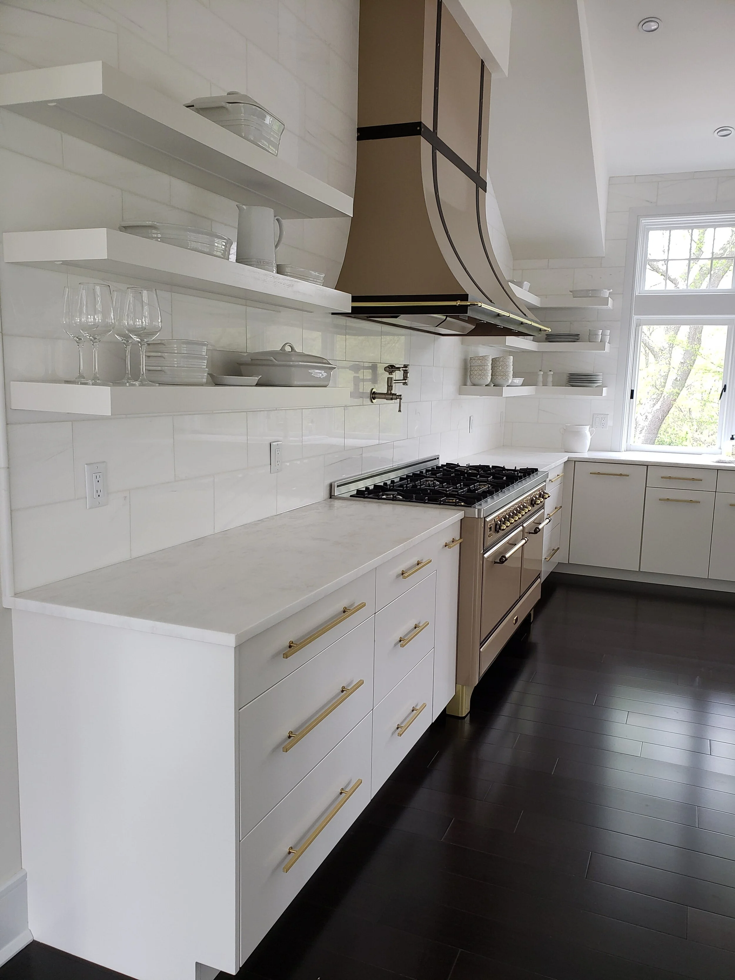 Modern kitchen with white cabinets, open shelves displaying glasses and dishes, a large window, dark hardwood floors, and a stainless steel stove with a range hood.