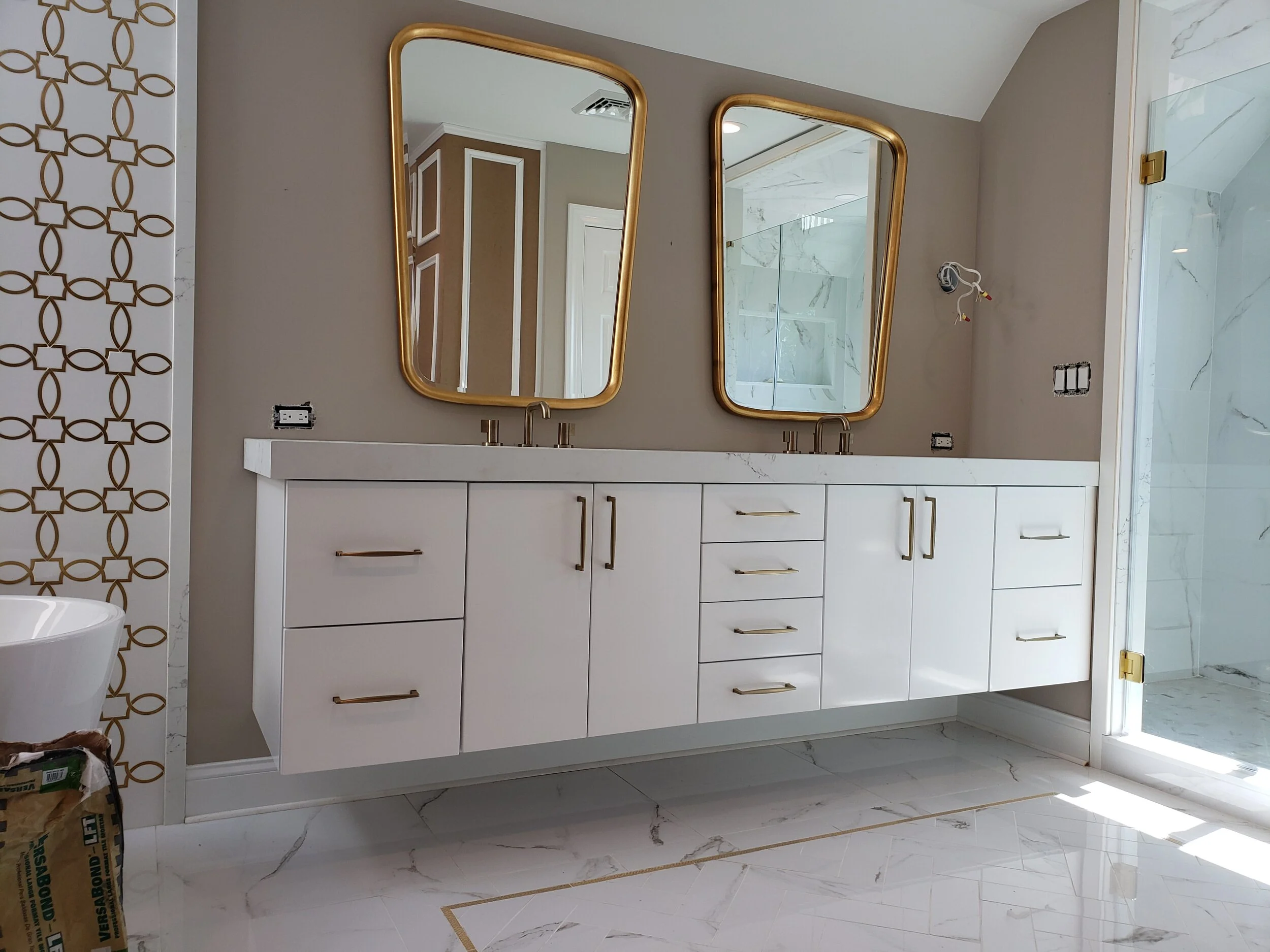 A modern white bathroom vanity with multiple drawers and cabinets, gold handles, two gold-framed mirrors above, set against a beige wall. There is a marble shower to the right and part of a white sink on the left.