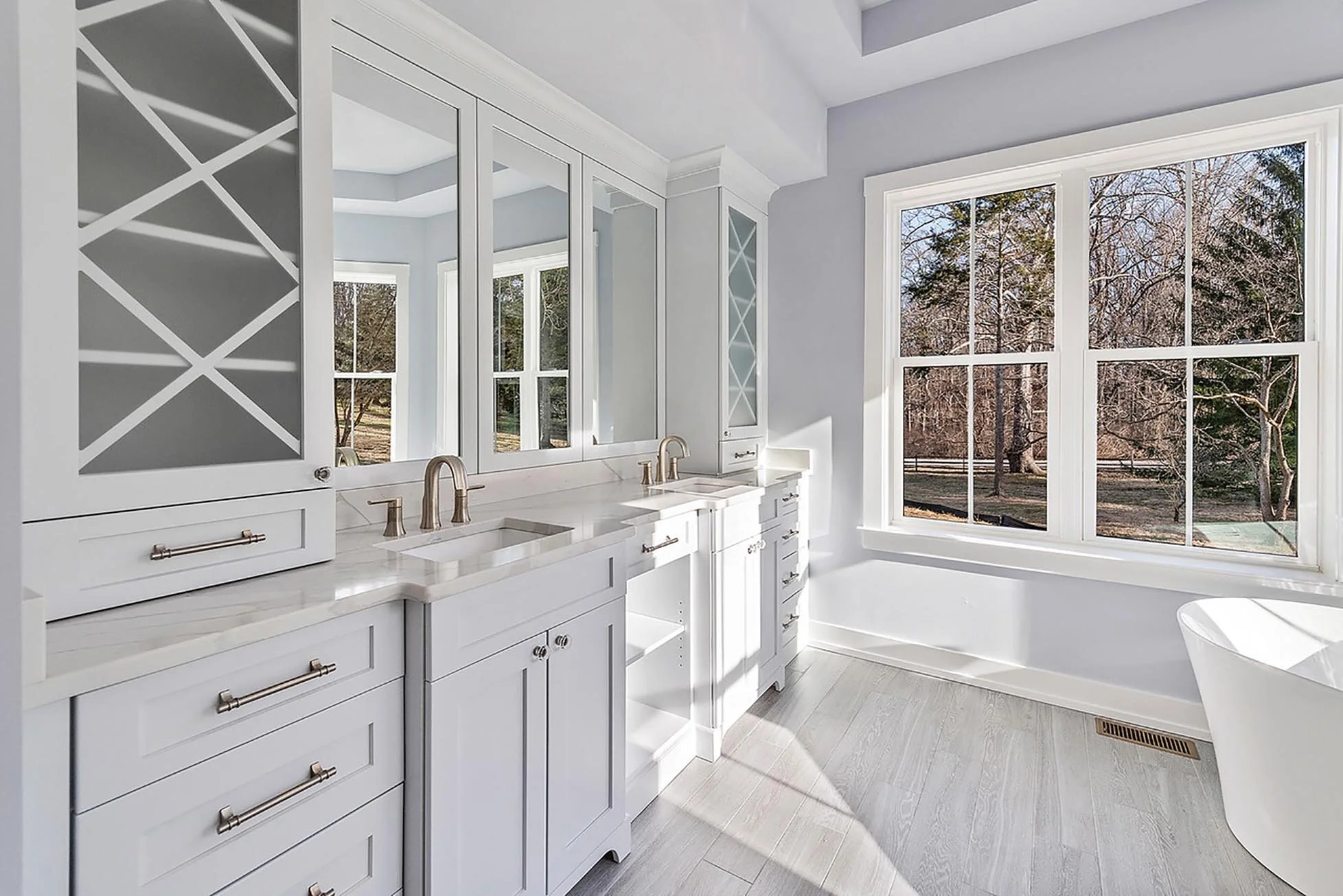 Bright bathroom with white cabinetry, double sinks, large window overlooking outdoor trees, and a bathtub.