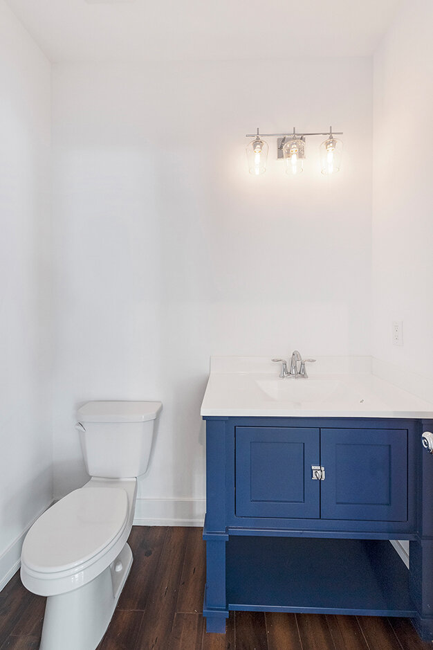 Bathroom with a white toilet on the left and a blue vanity with a white countertop and sink on the right, illuminated by a three-bulb light fixture.