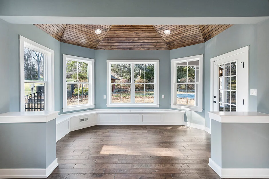 Bright sunlit room with large windows overlooking a backyard, wooden ceiling, and hardwood floors.