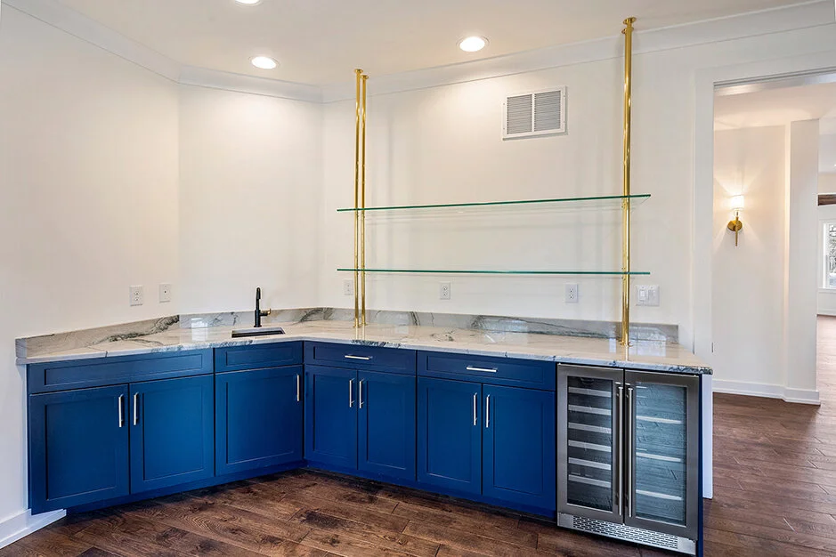 Kitchen bar area with blue cabinets, a marble countertop, glass shelves supported by gold poles, and a small wine cooler.