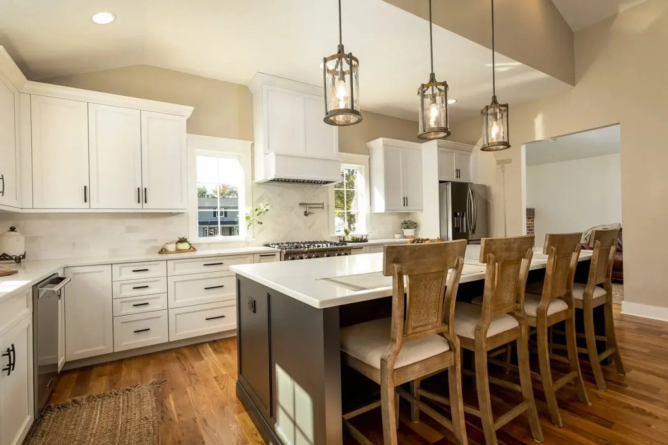 Modern kitchen with white cabinets, a large island with four wooden chairs, pendant lighting, stainless steel refrigerator, and hardwood floors.