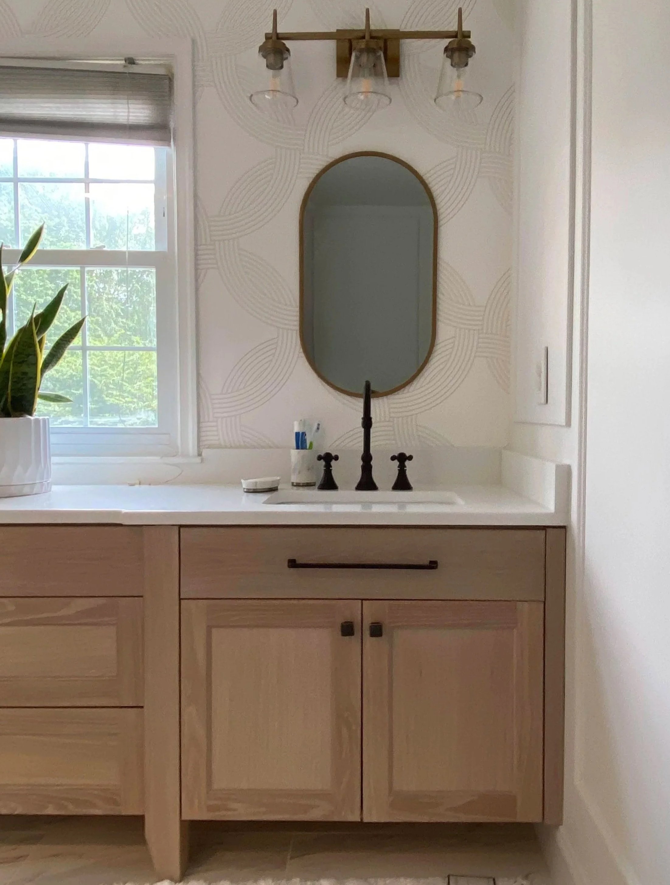Bathroom vanity with wooden cabinets, a white countertop, an oval mirror, a black faucet, a potted plant, and a window with sunlight.