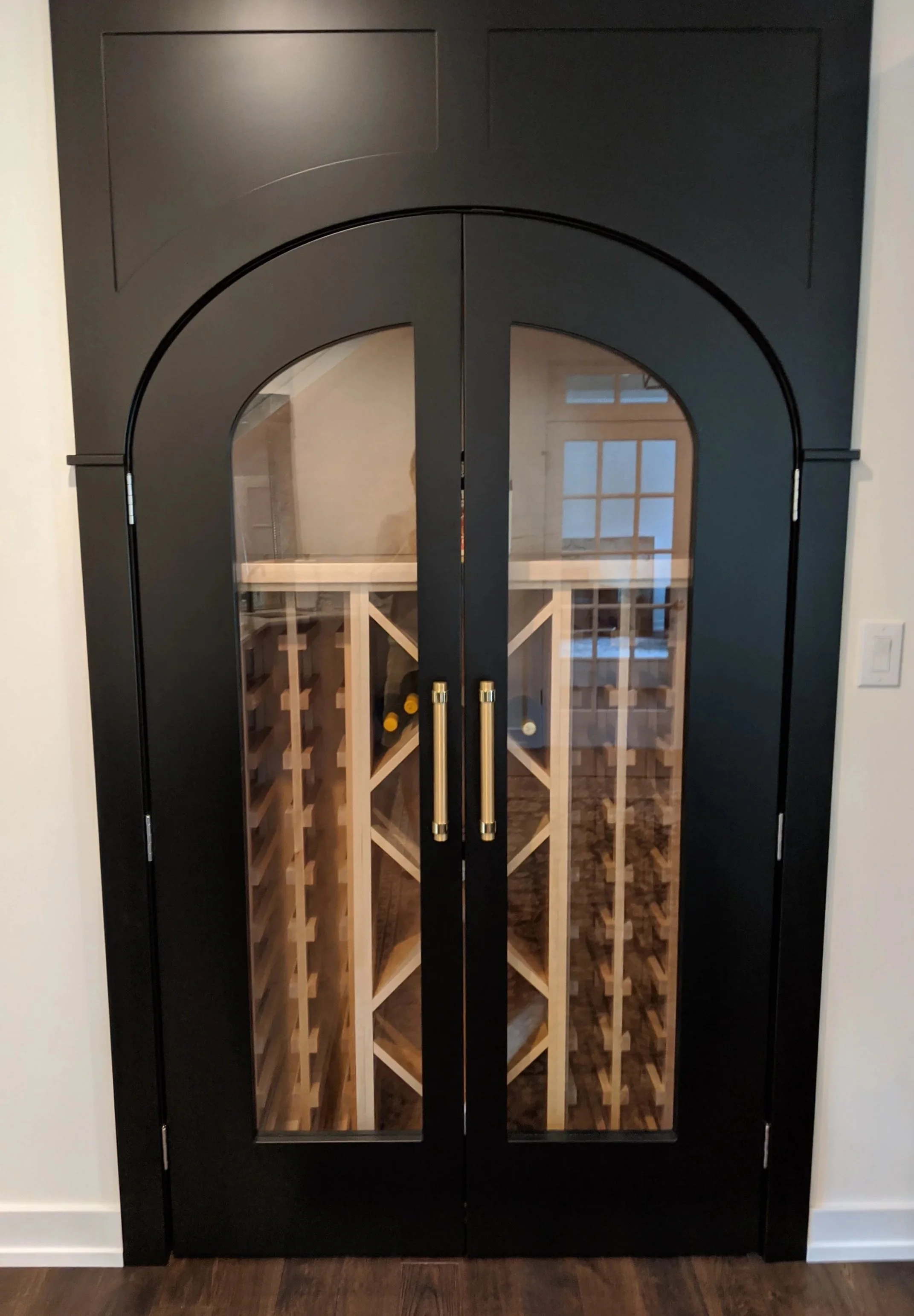 Black double doors with glass panels, opening to a wine cellar with wooden wine racks inside.