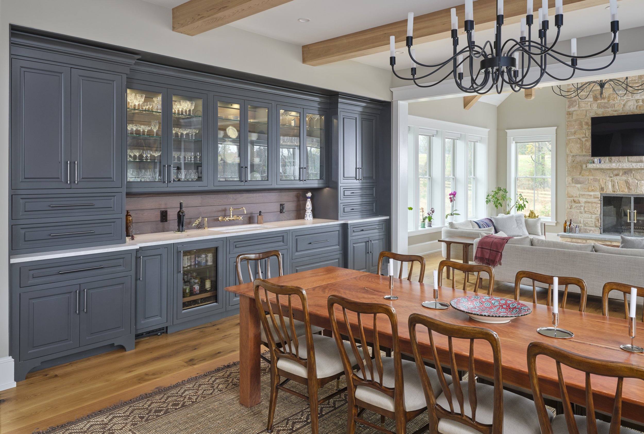 A dining and living area with a gray built-in cabinet, a wooden dining table with chairs, a chandelier, and a stone fireplace with a mounted TV.