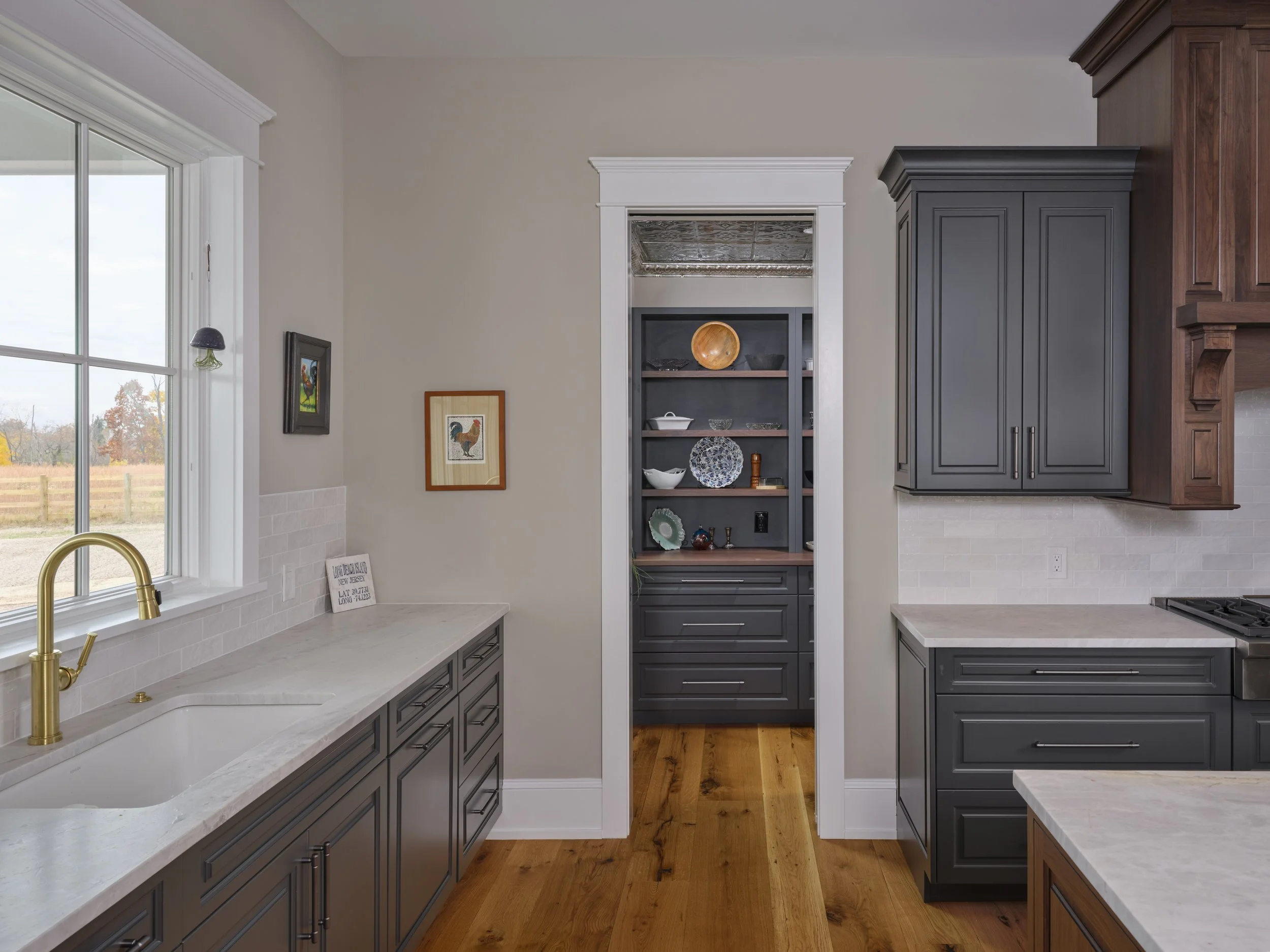 Views of a modern kitchen with gray cabinetry, white countertops, a window with a view outside, and a small built-in cabinet with dishes.