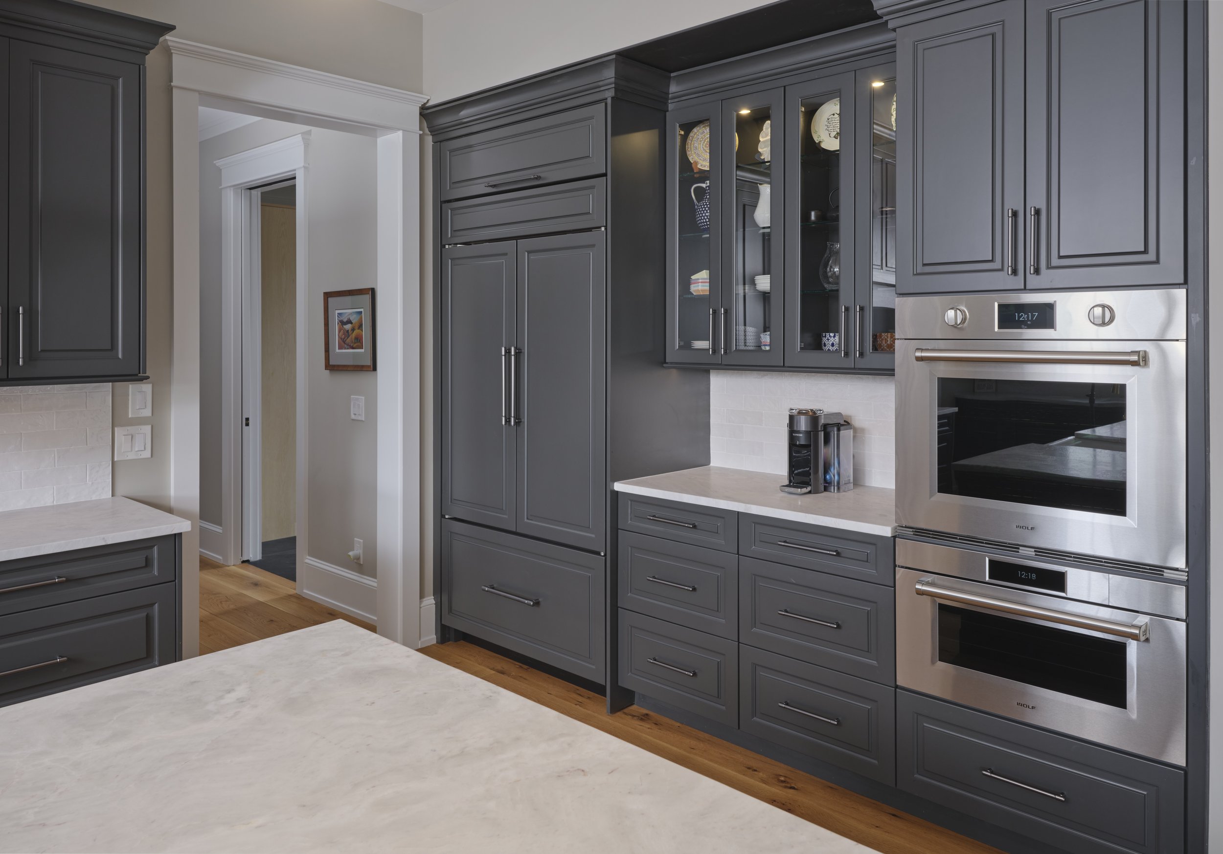 A modern kitchen with dark gray cabinets, white countertops, and stainless steel appliances, including a double oven, with a coffee maker on the counter.