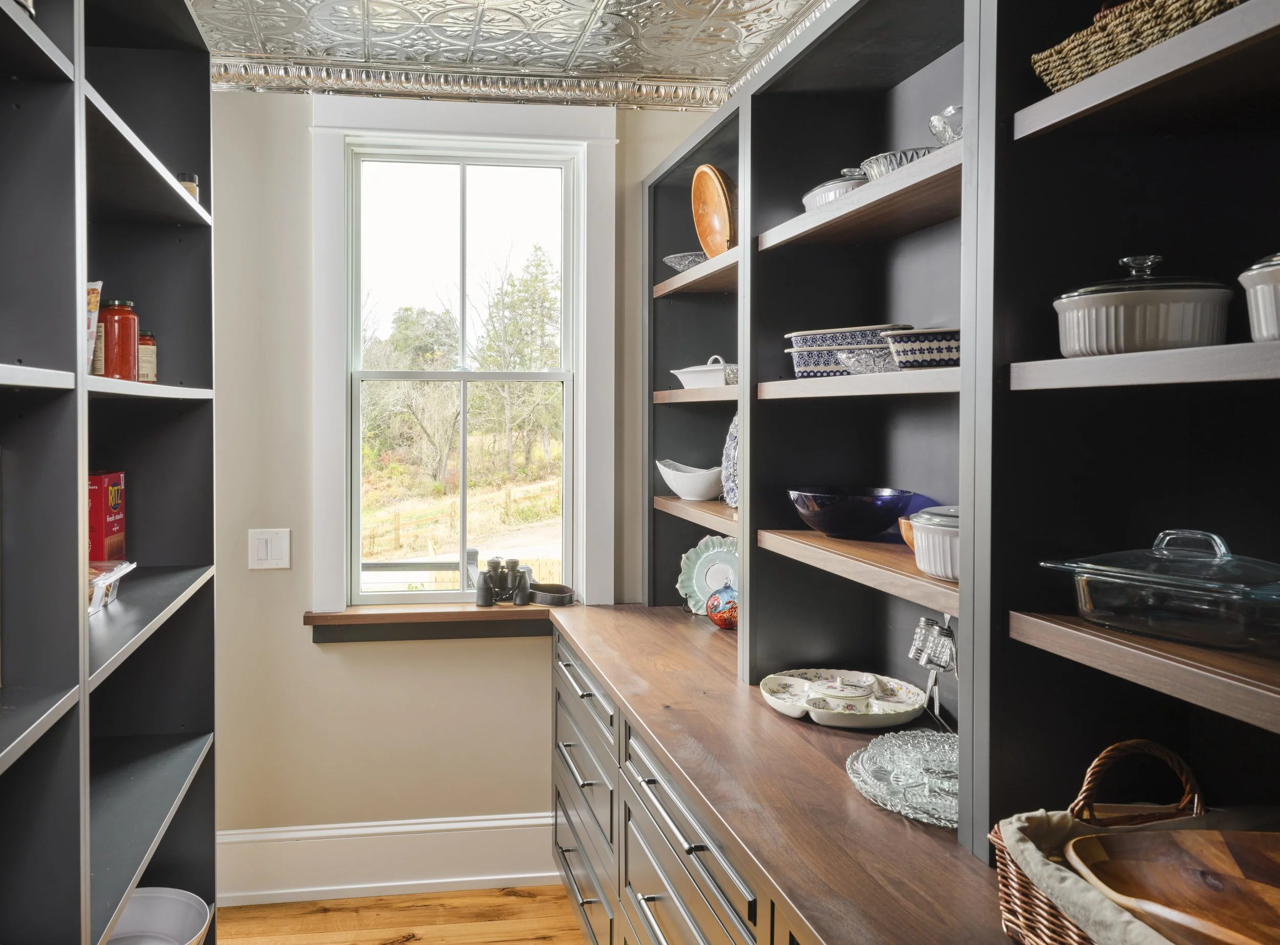 A kitchen pantry with open black shelving on both sides filled with dishes, bowls, and glassware. There is a wooden countertop surface with a window at the end showing an outdoor view with trees. The ceiling has decorative metallic tiles.