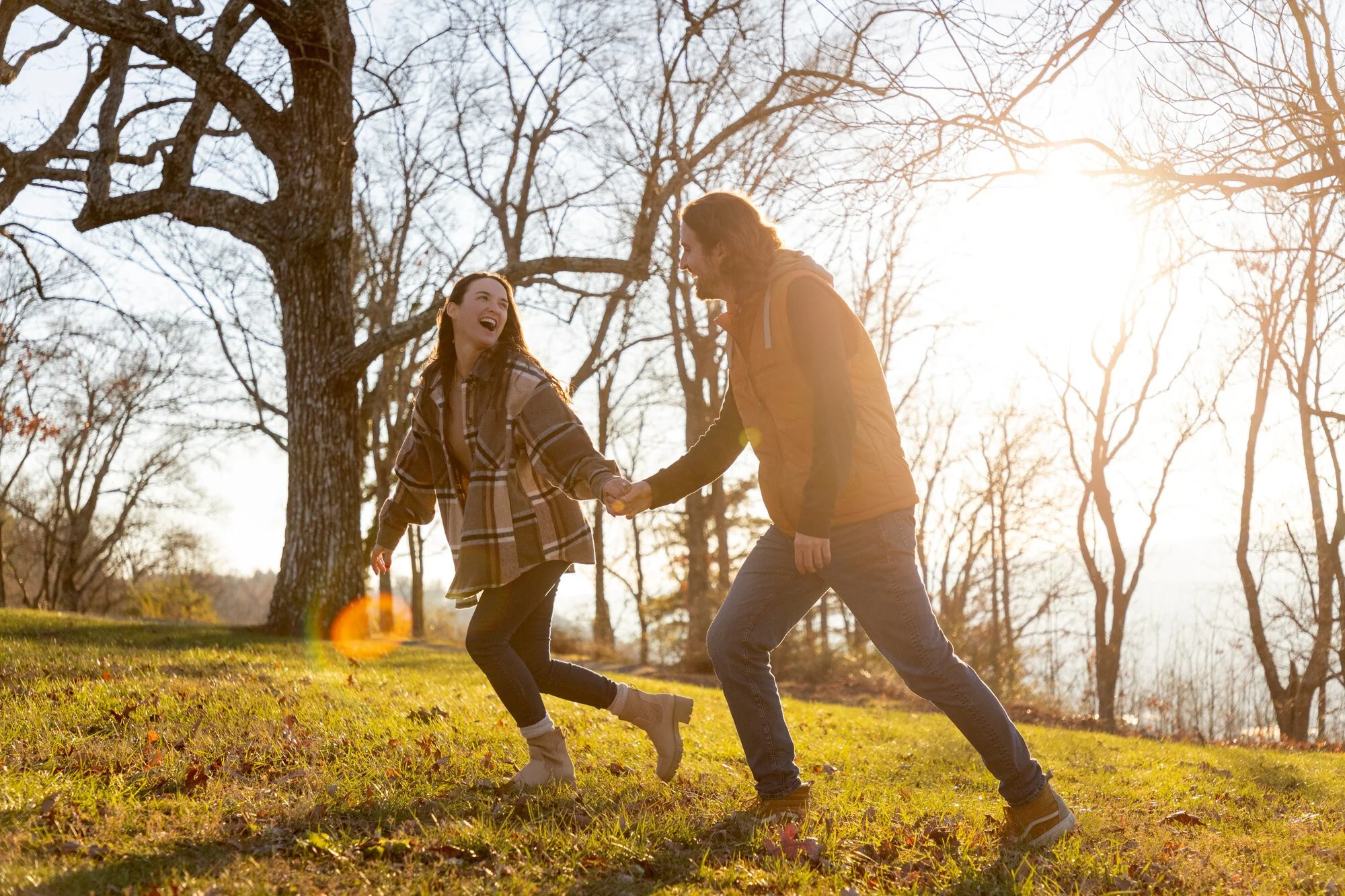 A young woman and a young man are holding hands and dancing in a park with bare trees during sunset.