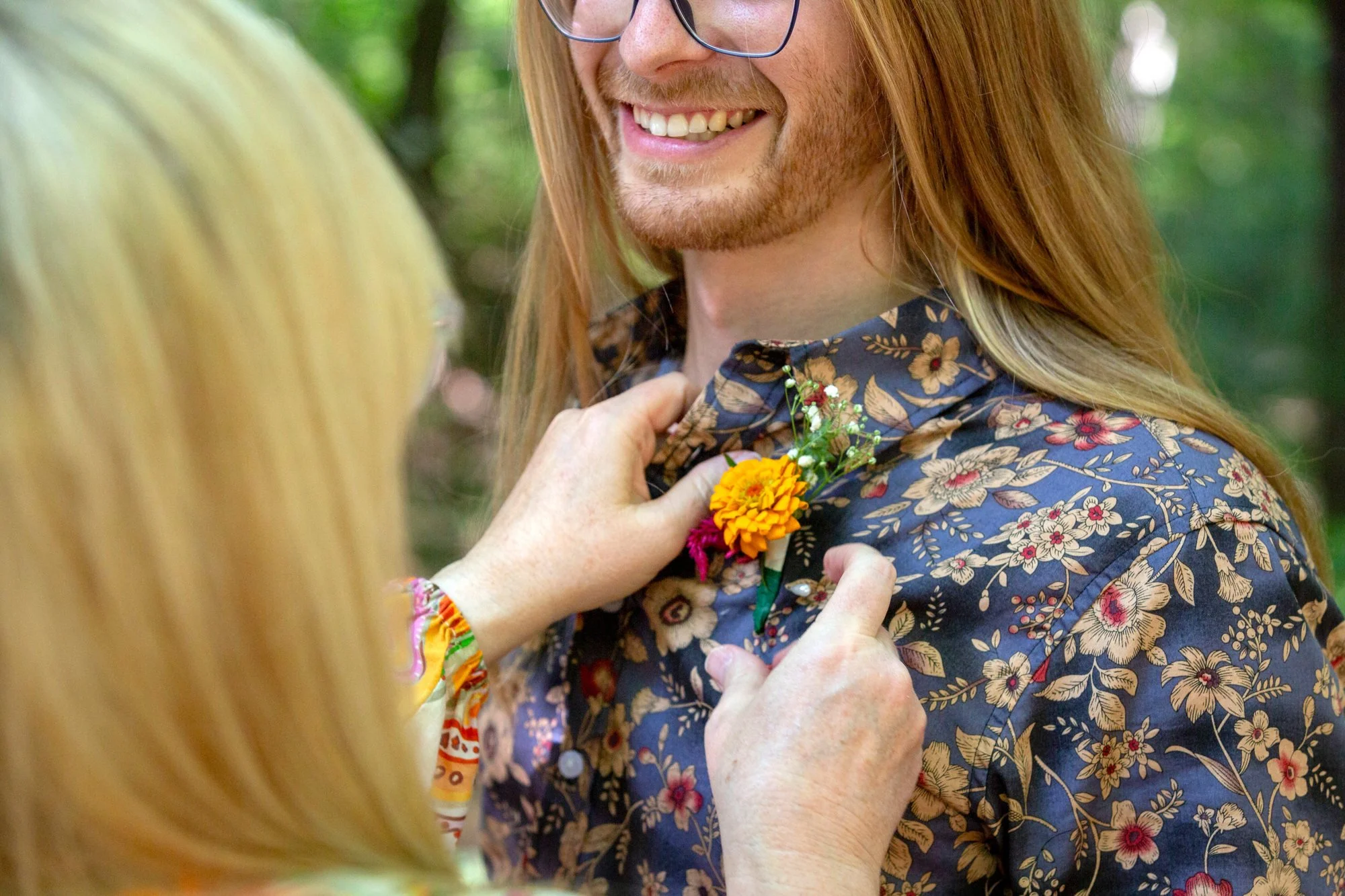 A person with long red hair and glasses is smiling as another person's hand pins a bouquet of colorful flowers onto their shirt, outdoors with green foliage background.