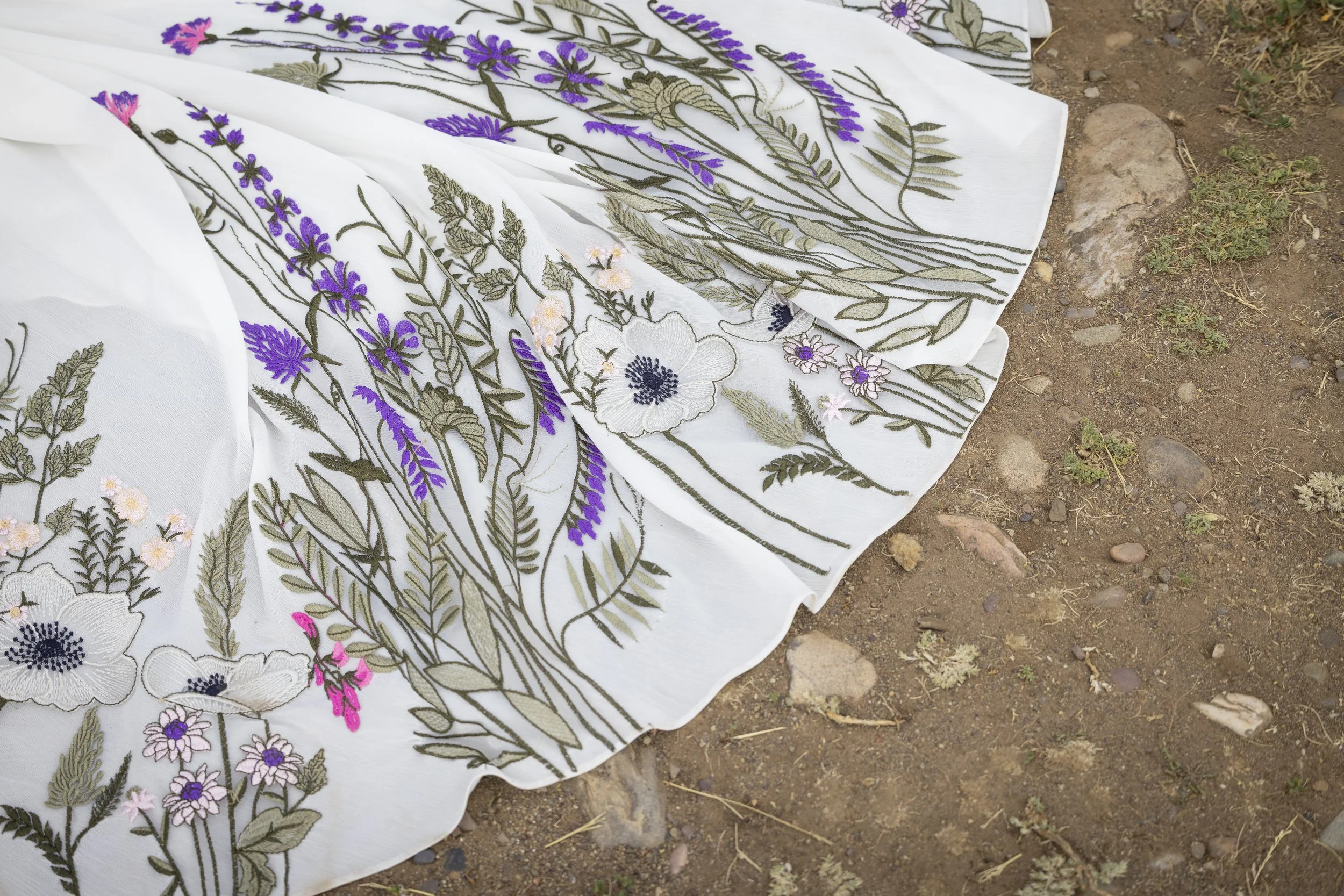 A white embroidered fabric with purple and pink flowers and green leaves, lying on dry dirt and small rocks.