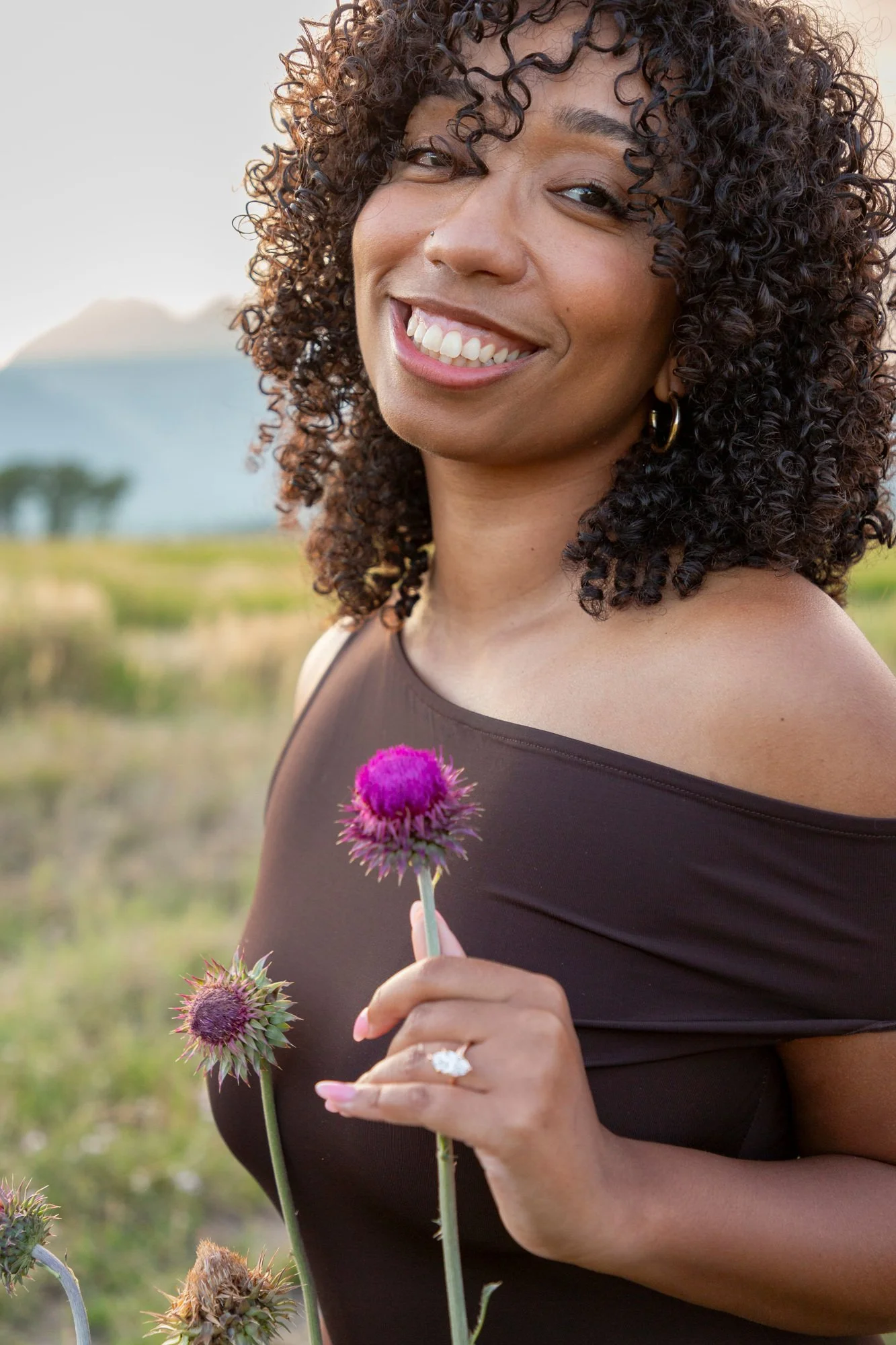 Smiling woman with curly hair holding purple thistle flowers outdoors