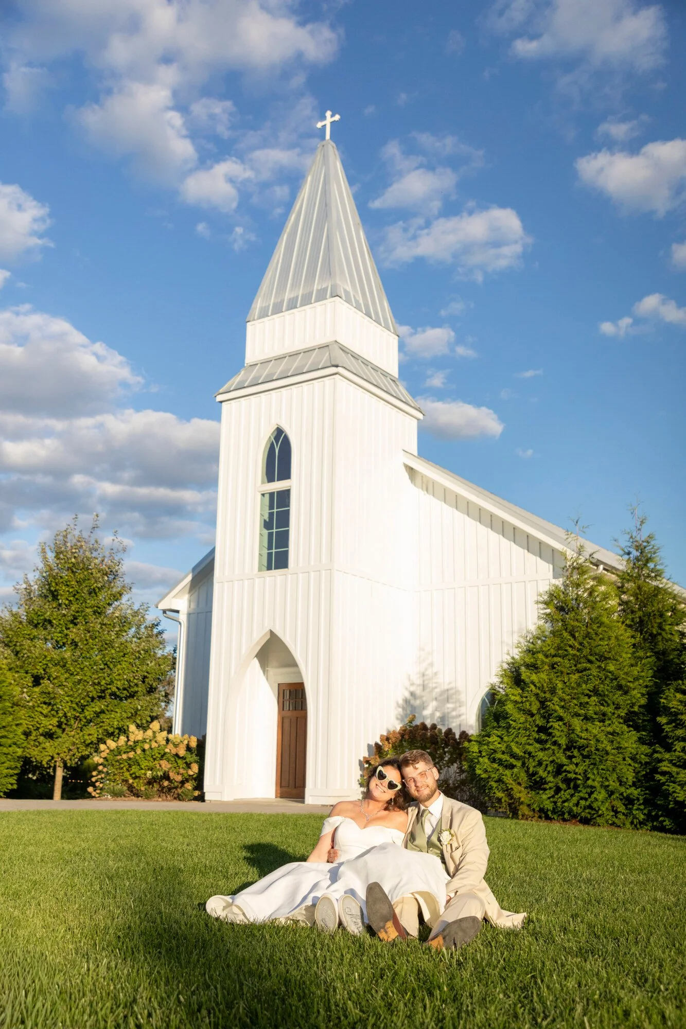 A newlywed couple sitting on the grass in front of a white church with a tall steeple topped with a cross. The bride is wearing a white wedding dress and sunglasses, and the groom is in a light-colored suit with sunglasses. They are smiling and embra