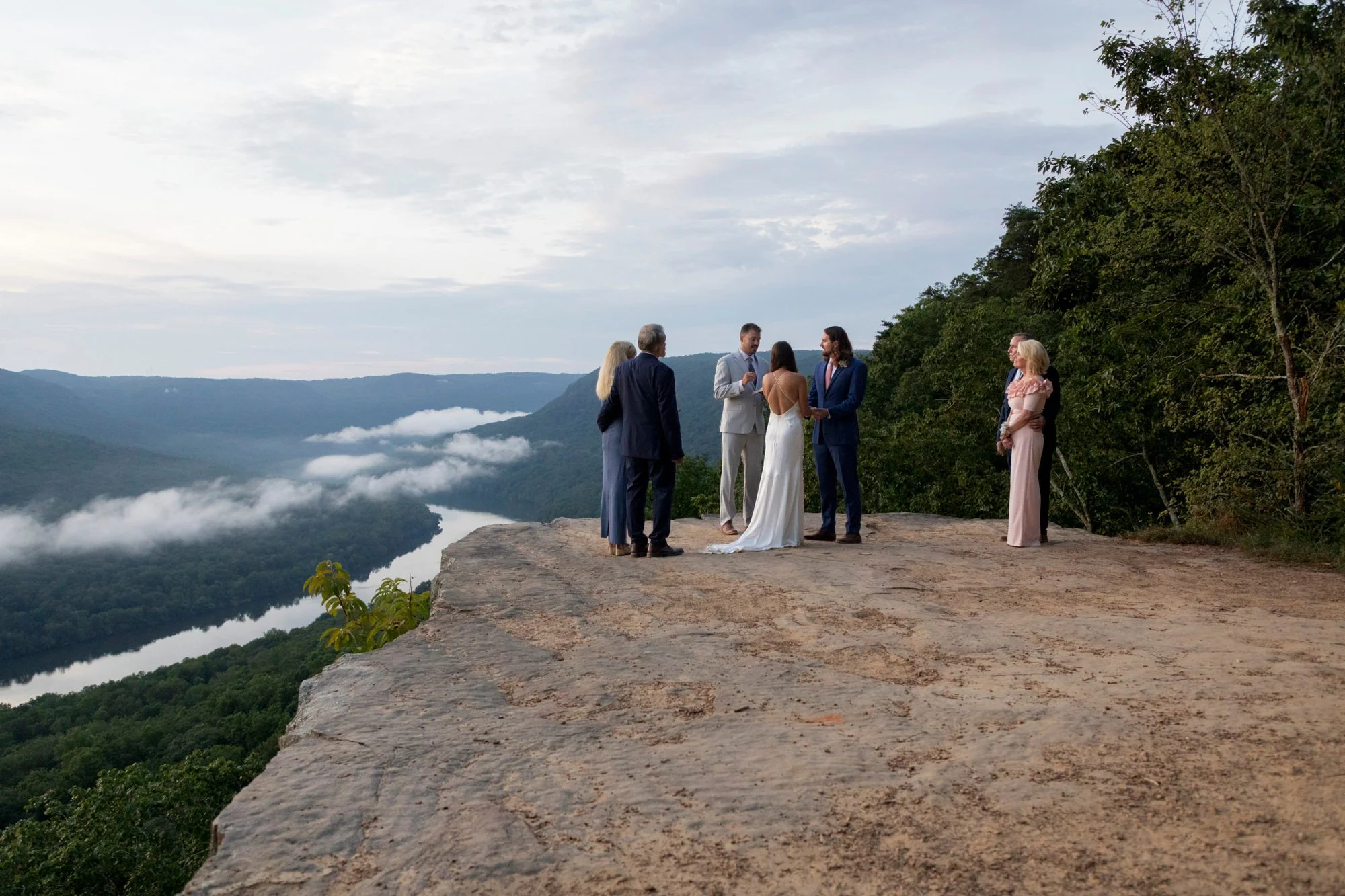 A wedding ceremony taking place on a rocky cliff overlooking a river in a mountainous landscape, with a few clouds in the sky and trees on the side.