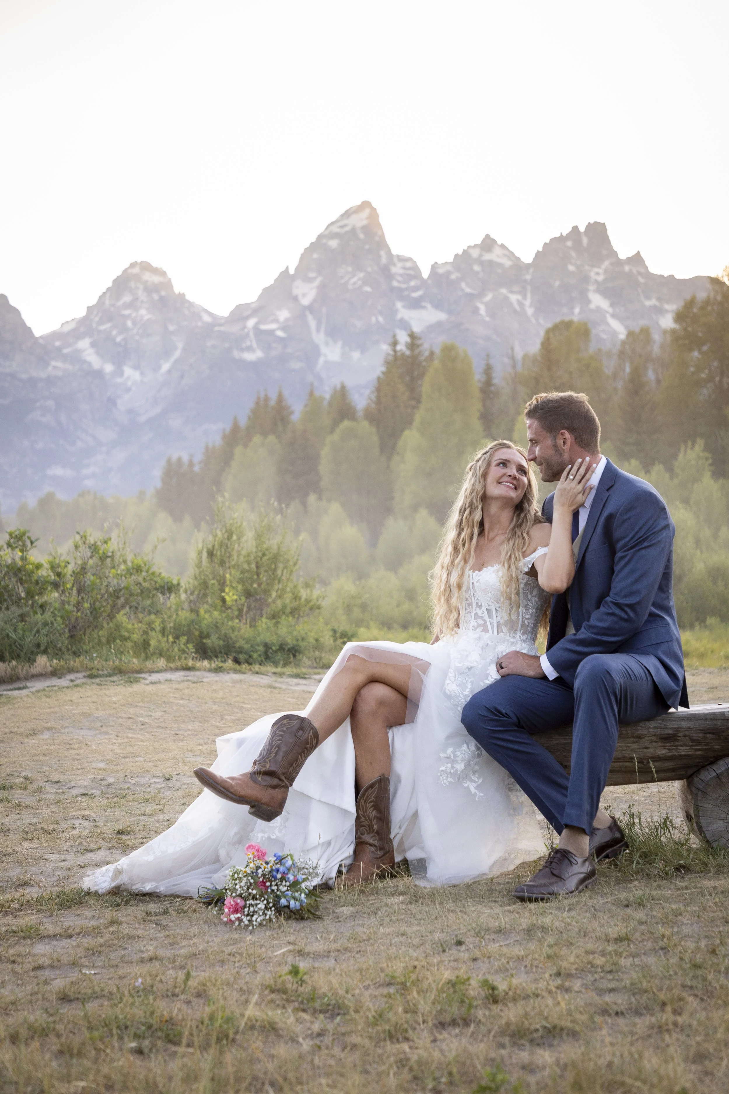 A couple dressed in wedding attire sitting outdoors on a log with a mountain range in the background during sunset.