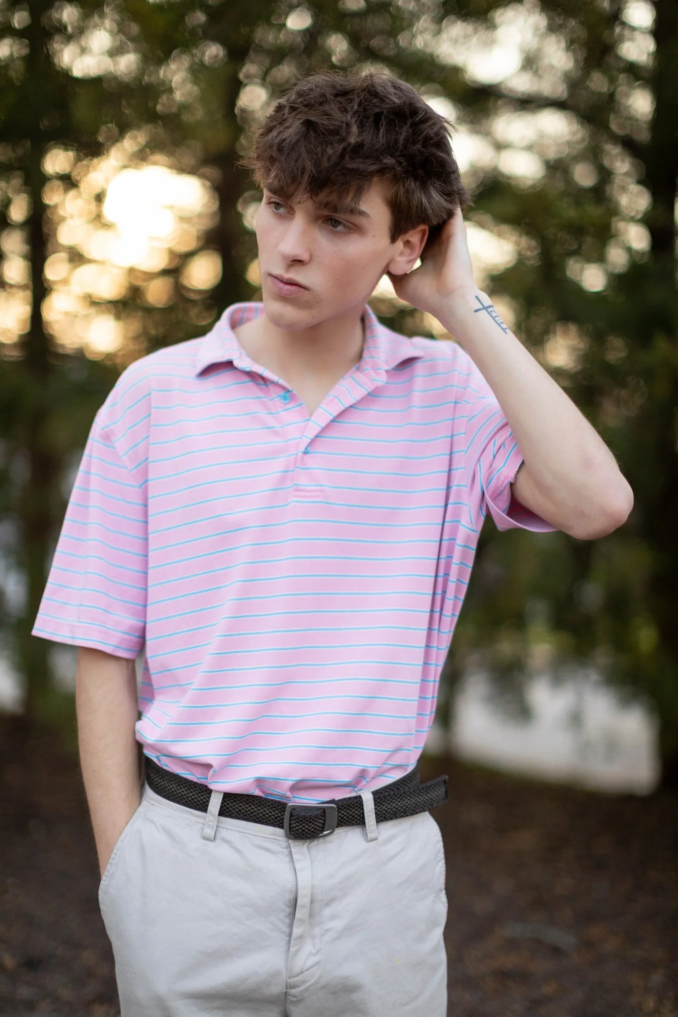 A young man with brown curly hair, wearing a pink striped polo shirt and light-colored pants, standing outdoors with trees and a bokeh background, touching his head with one hand and looking to the side.