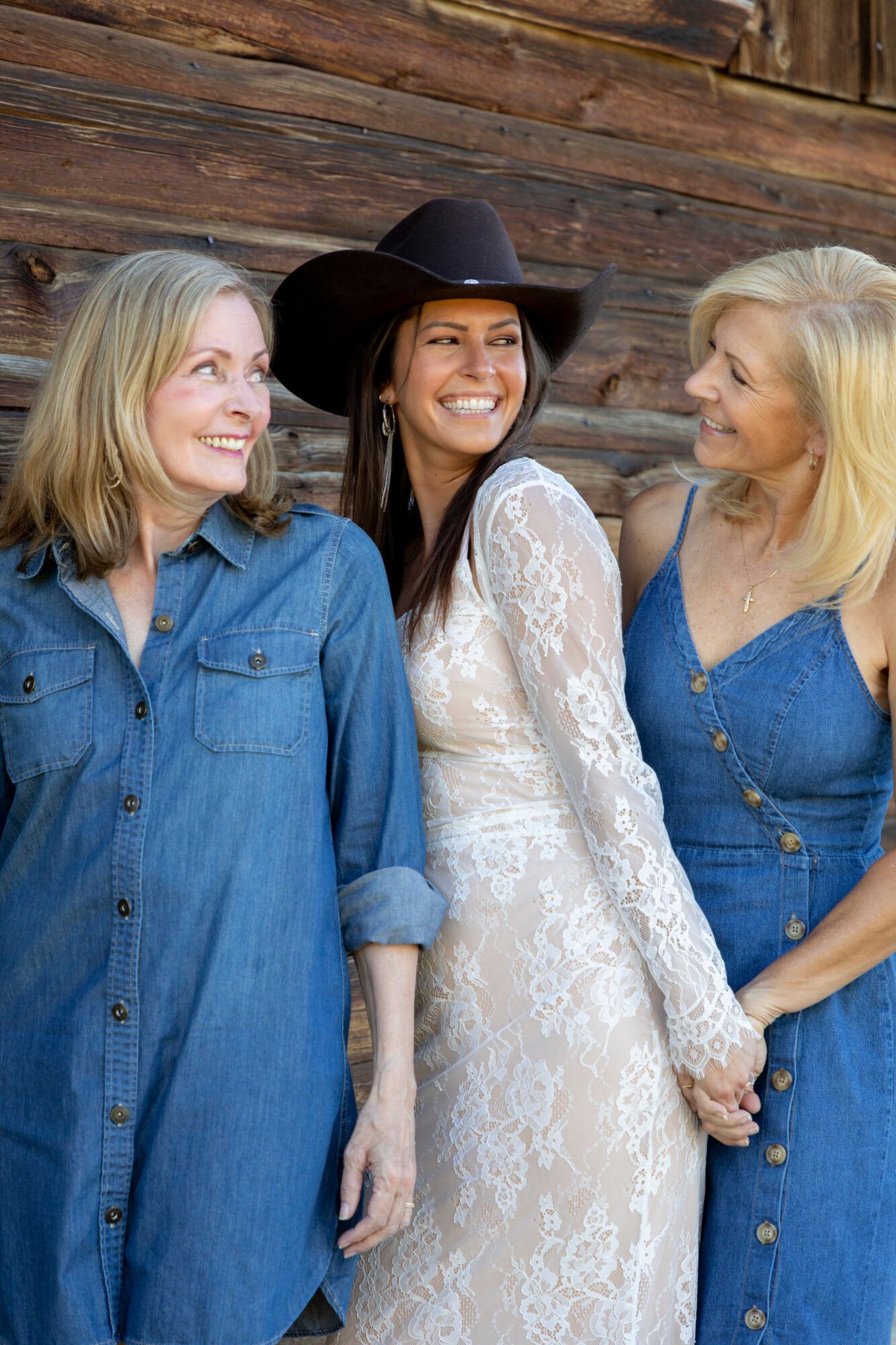Three women standing together, holding hands and smiling. The woman in the middle wears a black cowboy hat and a white lace dress, while the women on her sides wear denim dresses. They are standing against a wooden wall.