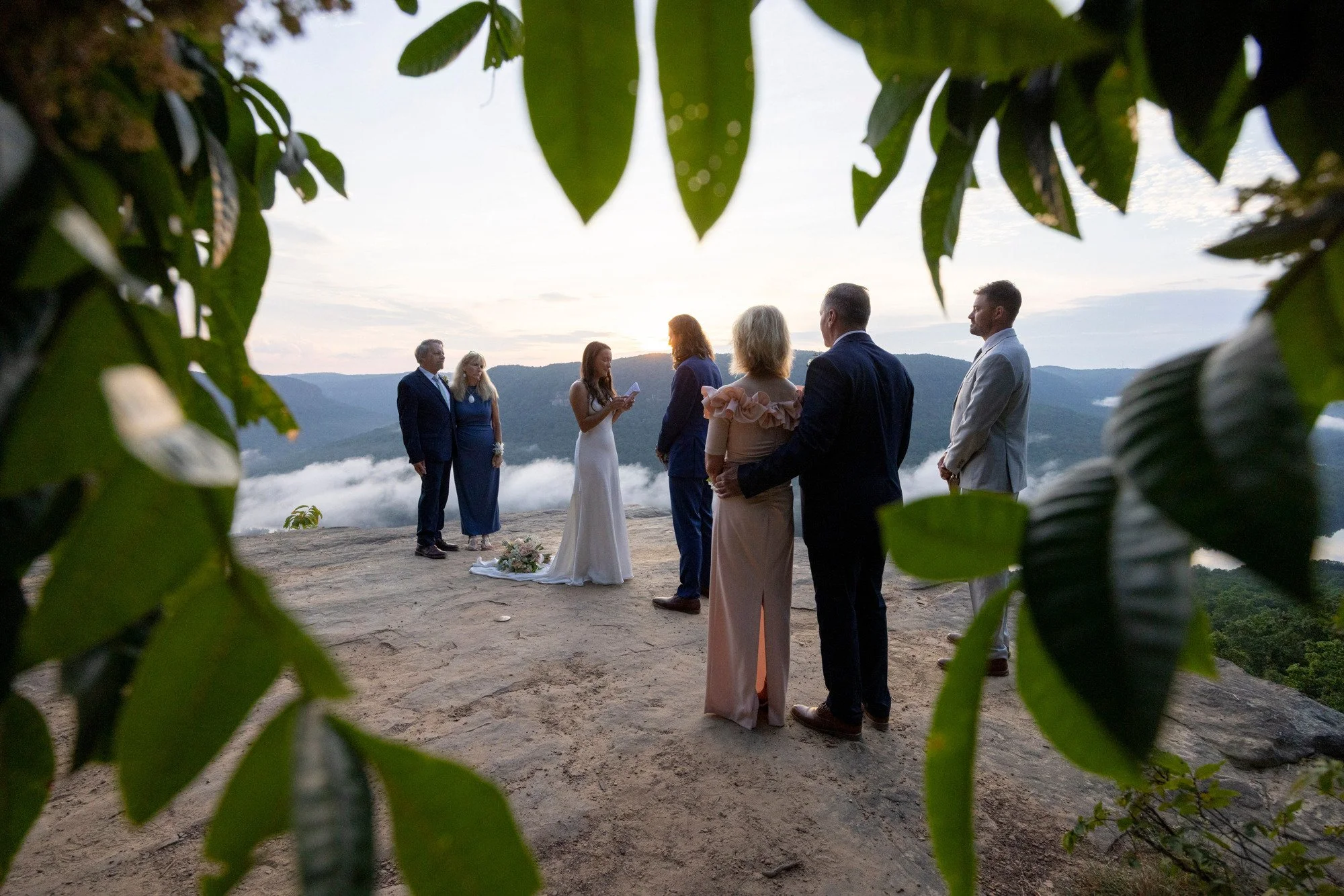 A wedding ceremony taking place outdoors on a mountain top with six people gathered around a bride and groom. The scene is framed by green leaves in the foreground, and the background features mountain ridges and a cloudy sky at sunset.