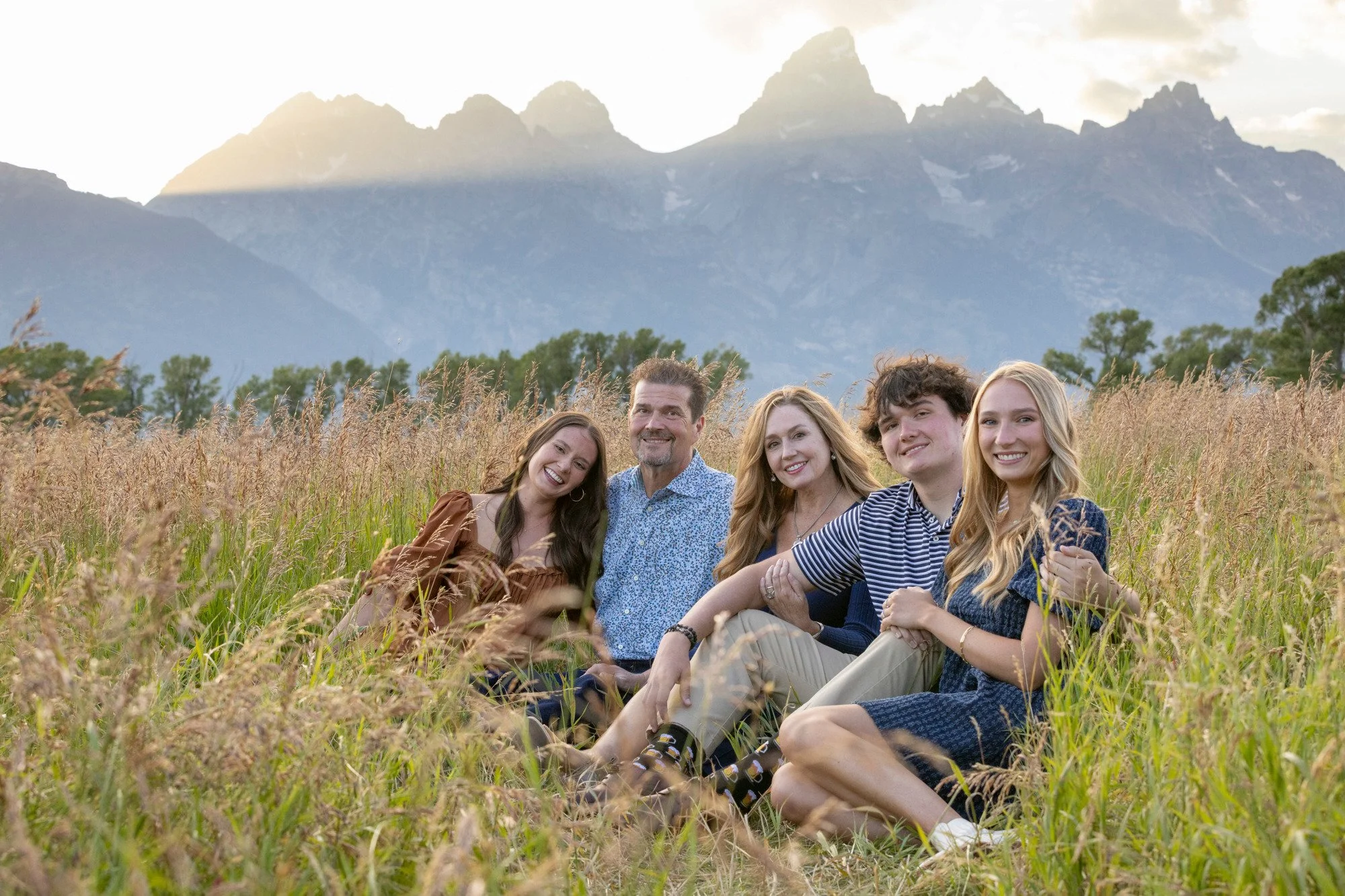 A family of five sitting in a grassy field with mountains in the background during sunset. The family members are smiling and dressed casually.