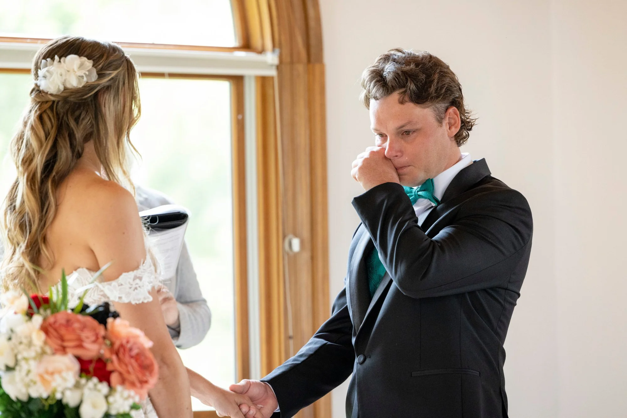 A man in a tuxedo with a teal bow tie, tears in his eyes, holding hands with a woman who is wearing a wedding dress, in a room with wooden frame windows.