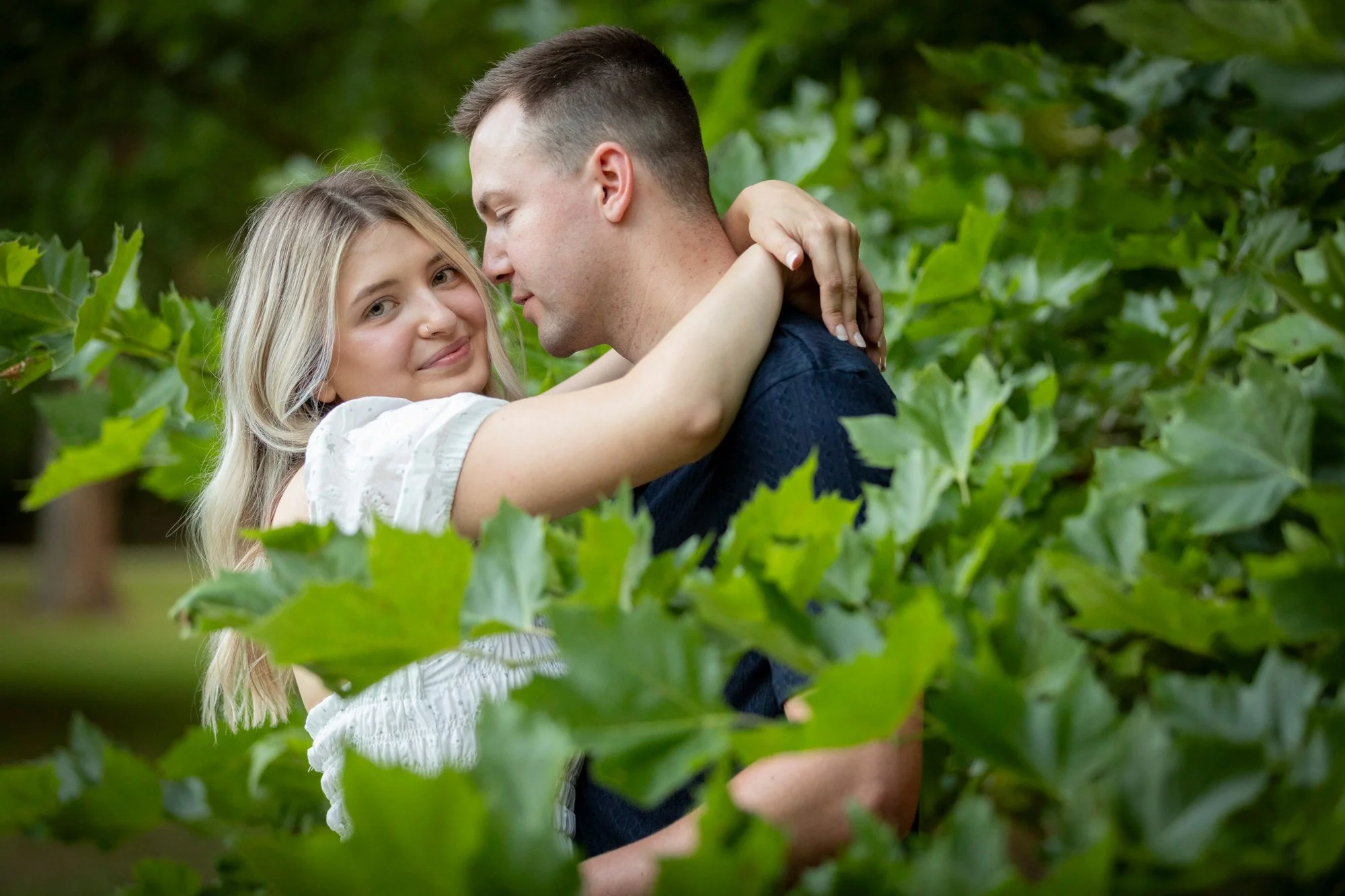A young couple embracing in a green outdoor setting, surrounded by leafy trees.