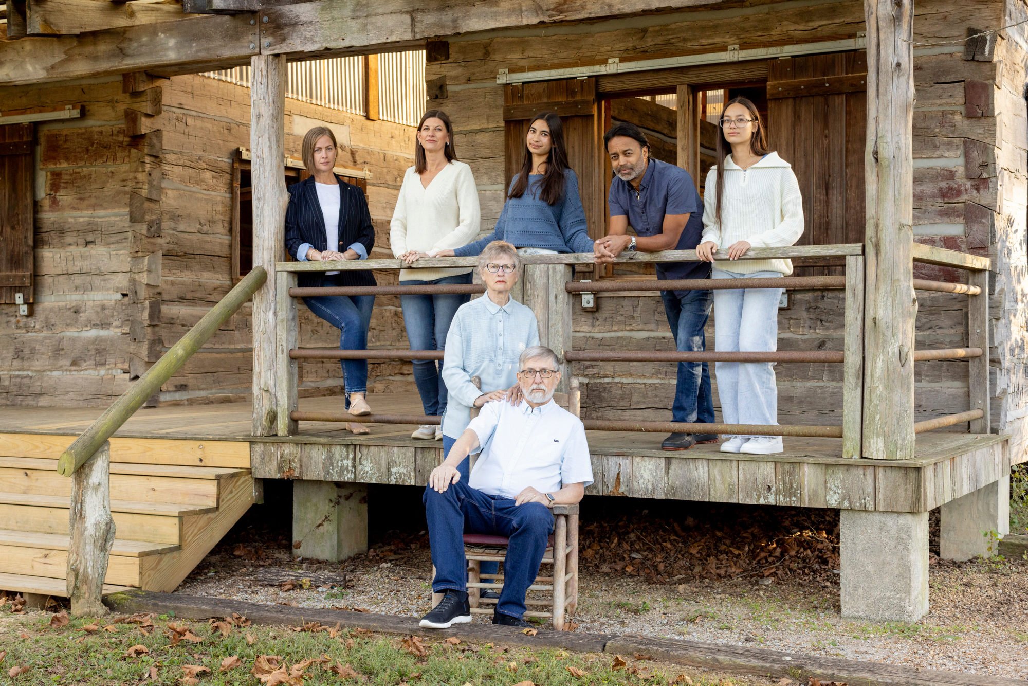 Group of seven diverse people, including women and men of various ages and ethnicities, posing on a wooden porch of a rustic cabin during daytime.