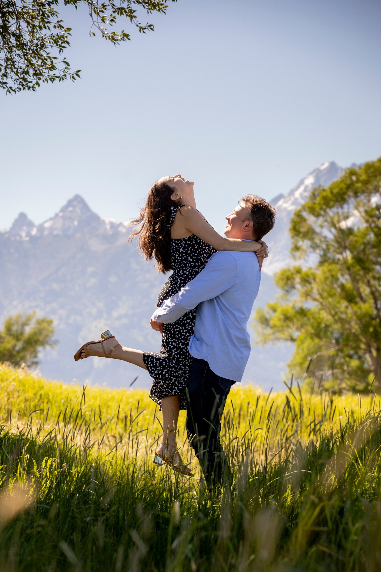 A man lifting a woman in a field with mountains and trees in the background.