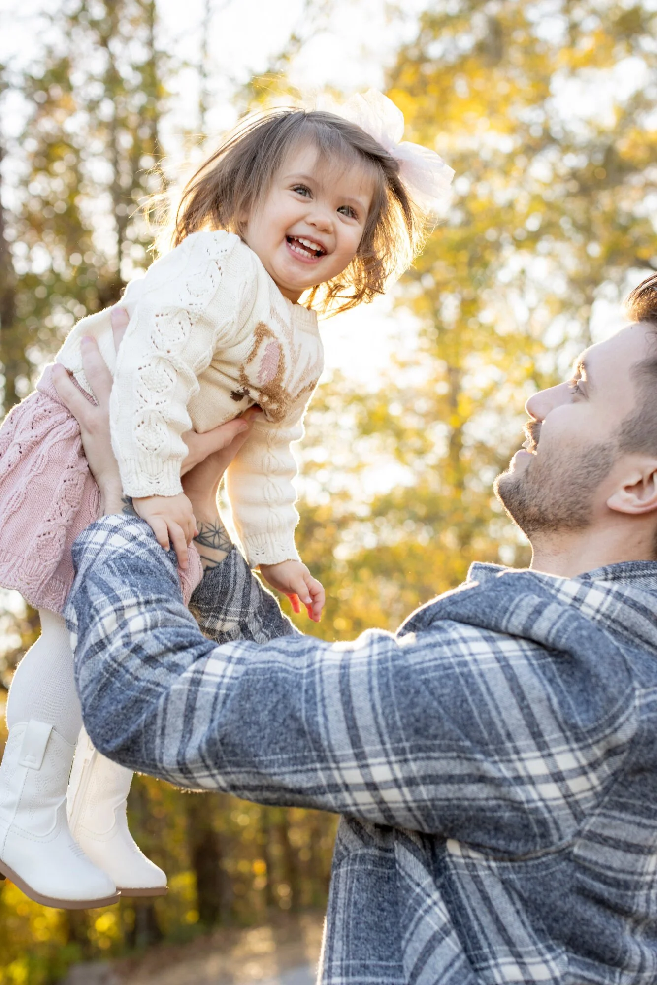 A man lifting a young girl in an outdoor park during autumn, with trees and golden leaves in the background.