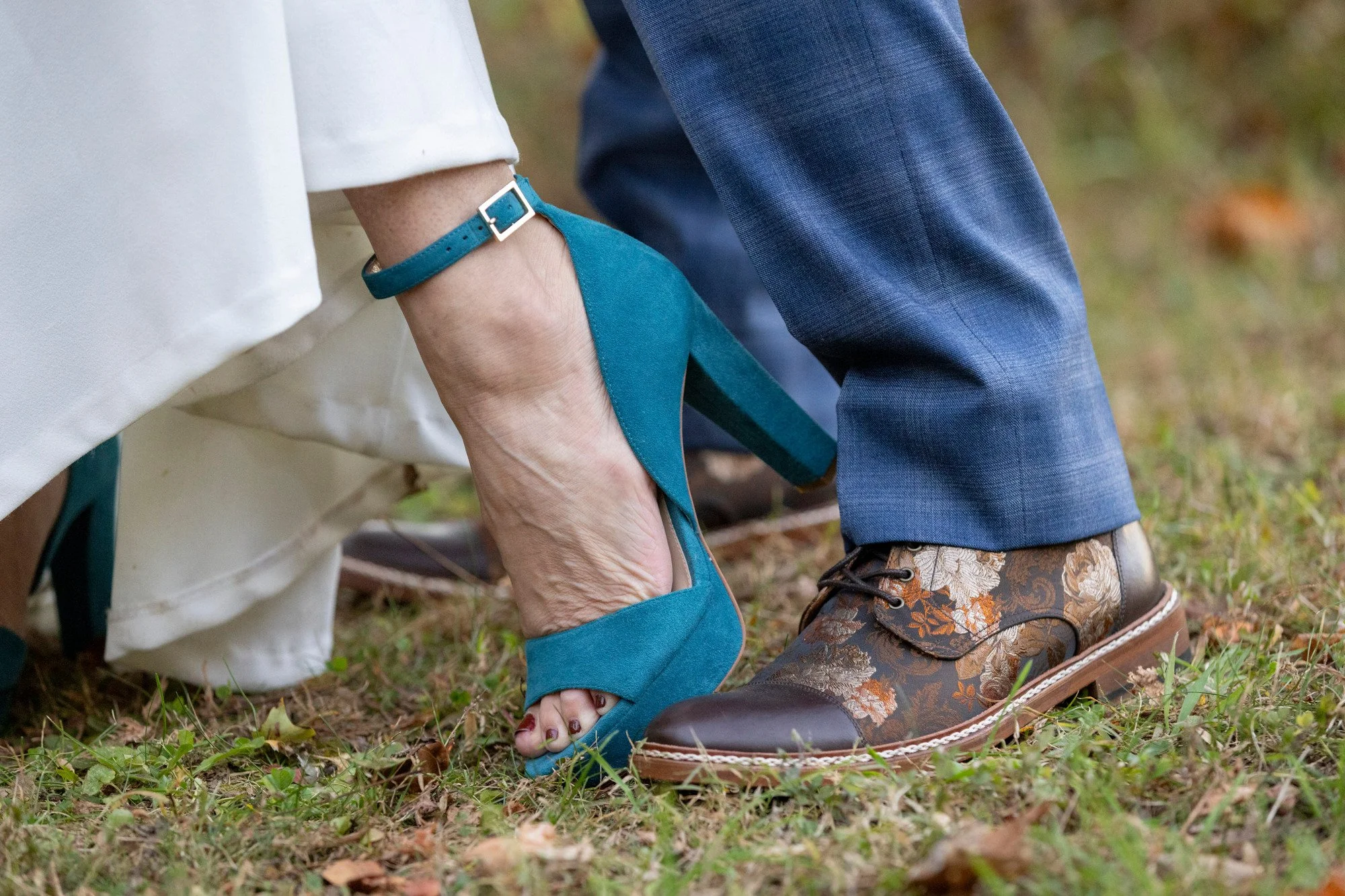 Close-up of a person wearing teal high-heeled shoes, a teal watch, and light-colored pants, kneeling outdoors on grass with another person wearing dark blue pants and floral-patterned shoes standing nearby.