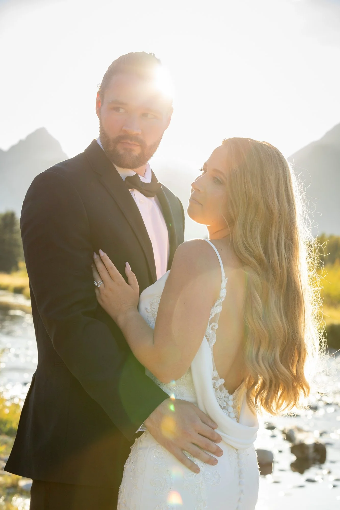 A couple in wedding attire standing outdoors near a body of water with mountains in the background, backlit by the sun, creating a bright, golden glow.