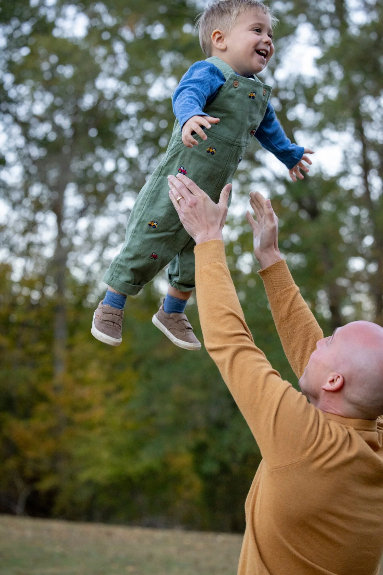 A man lifts a young boy into the air outdoors, with trees in the background, during daytime.