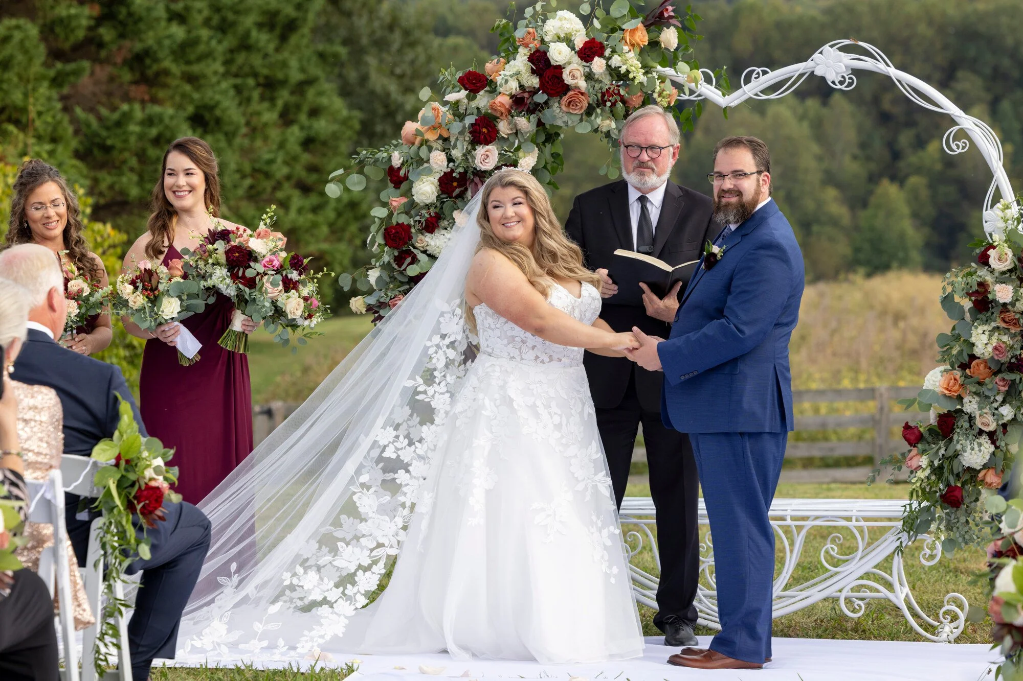 A wedding ceremony outdoors with a bride and groom holding hands and facing each other beneath a floral arch. The bride is wearing a white wedding dress with lace details and a long veil, and the groom is dressed in a blue suit with a boutonniere. Th