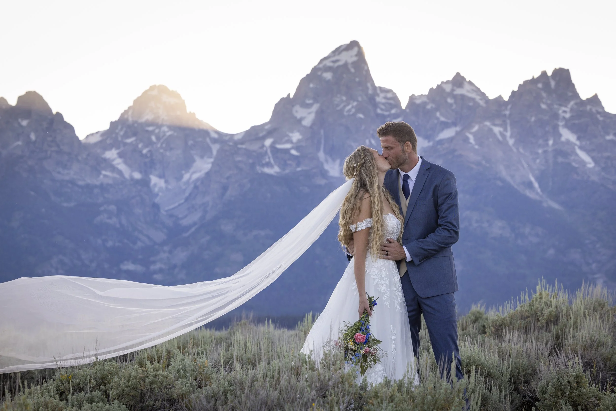 A bride and groom sharing a kiss outdoors at sunset with mountain peaks in the background, the bride holding a bouquet.