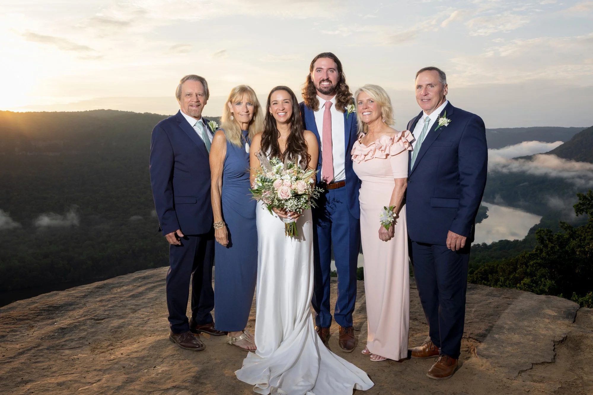 A group of six people, including a bride holding a bouquet, standing outdoors on a rock with a scenic mountainous background at sunset.