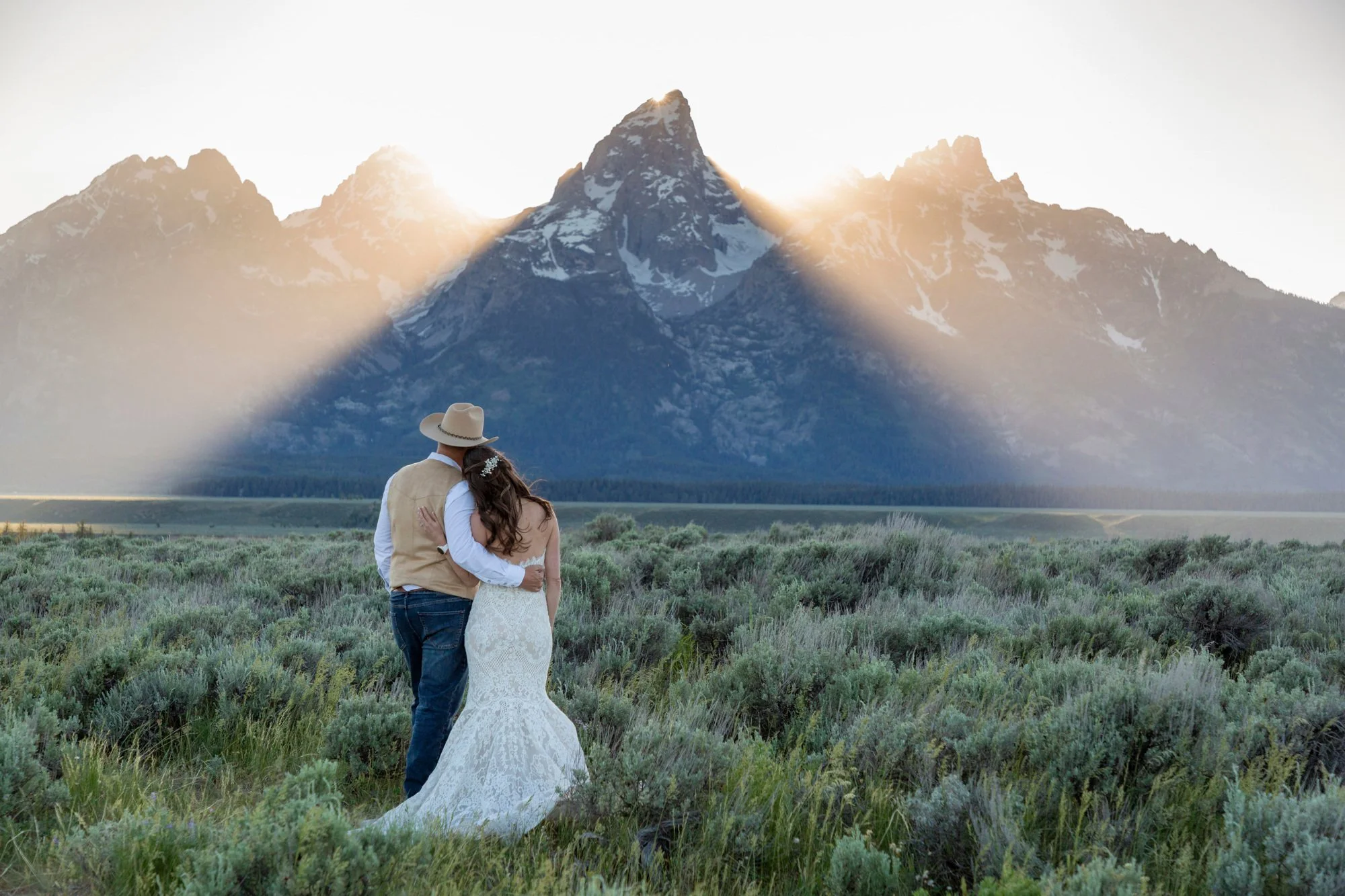 A couple, with the woman in a white wedding dress and the man in a beige vest and cowboy hat, embracing in front of a mountain backdrop during sunset.