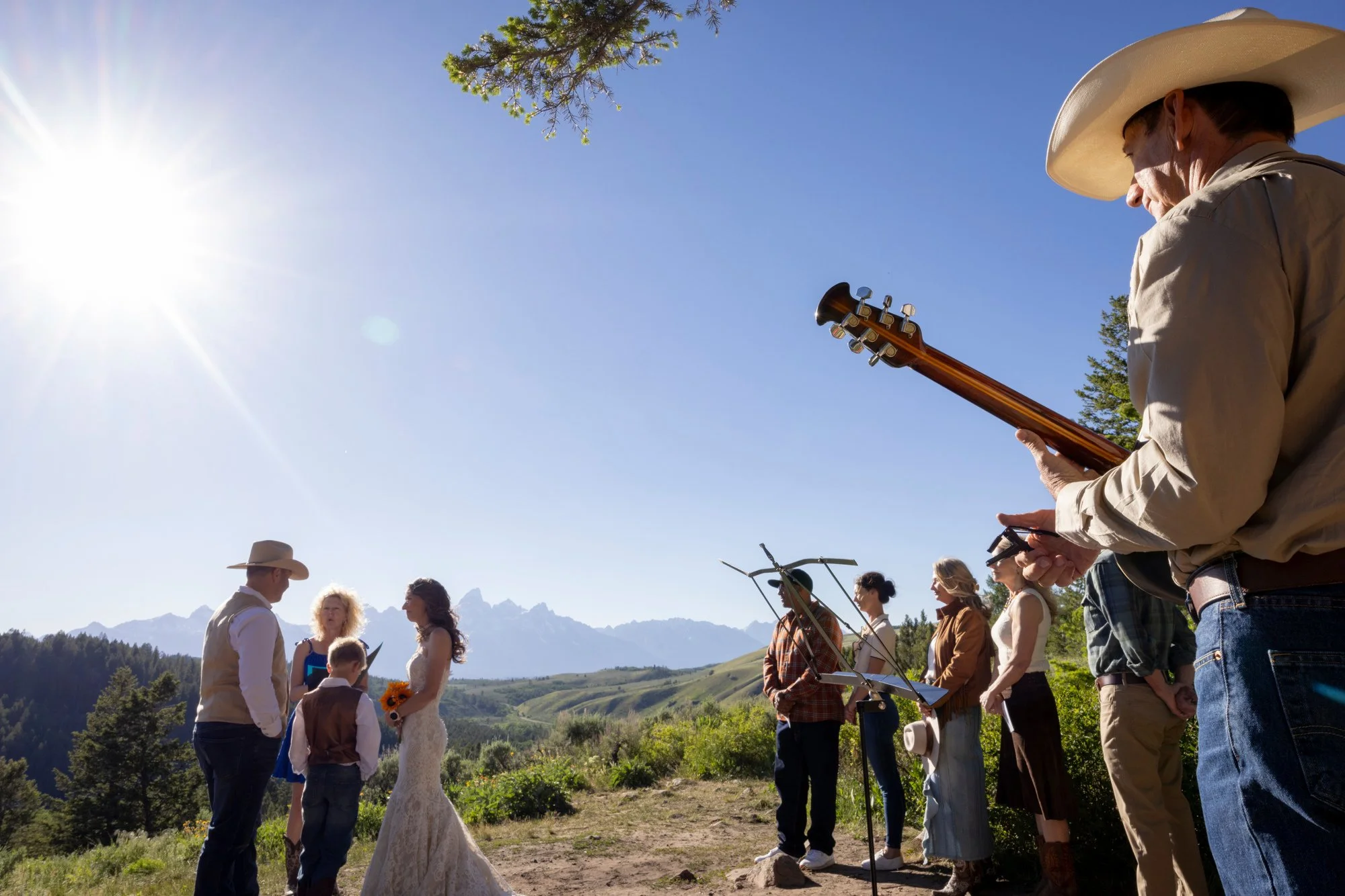 A bride and groom exchanging vows outdoors on a sunny day, surrounded by friends and family, with a musician playing guitar nearby and mountains in the background.