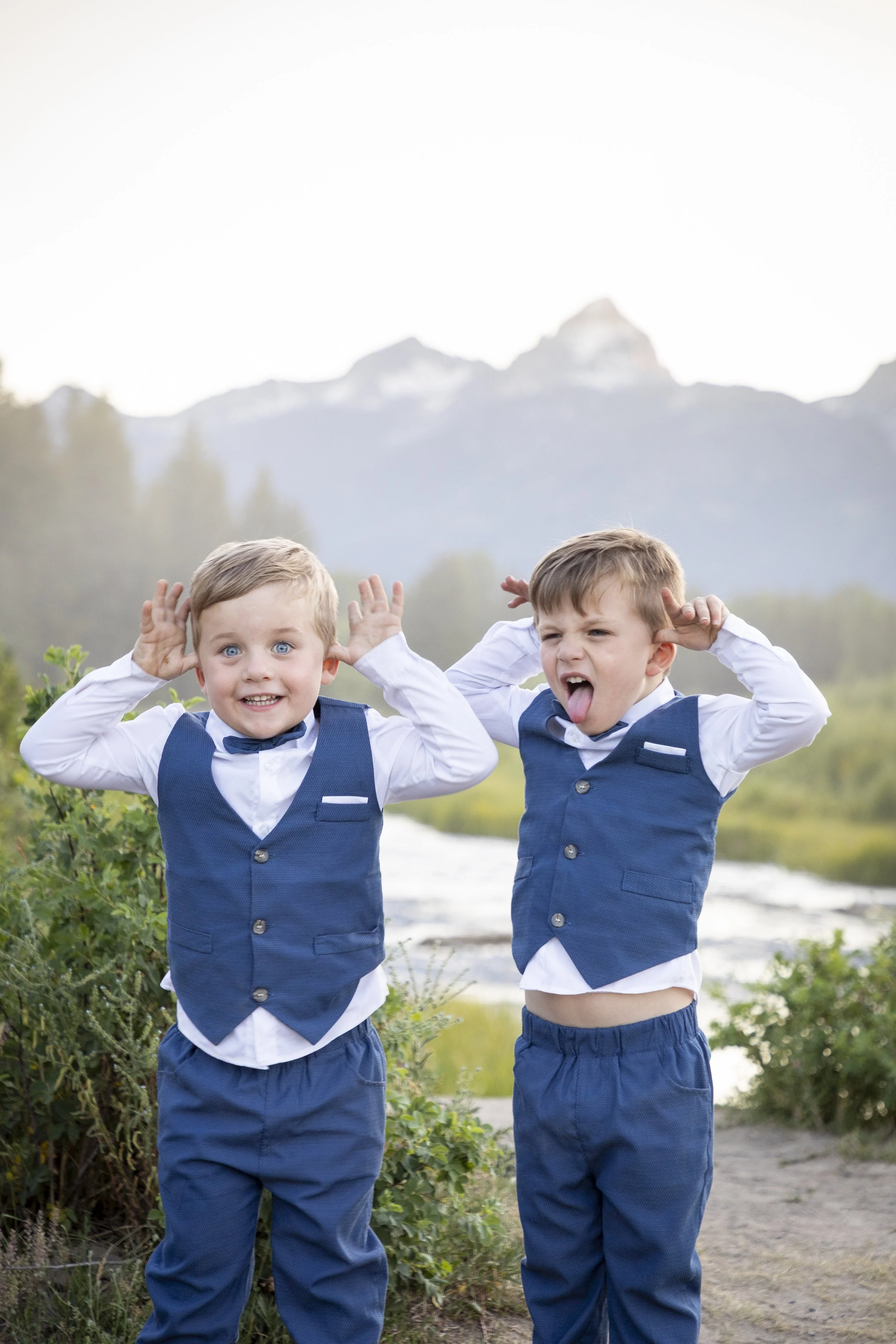 Two young boys standing outdoors near a river with mountains in the background, wearing matching blue vests and white shirts, making playful faces.