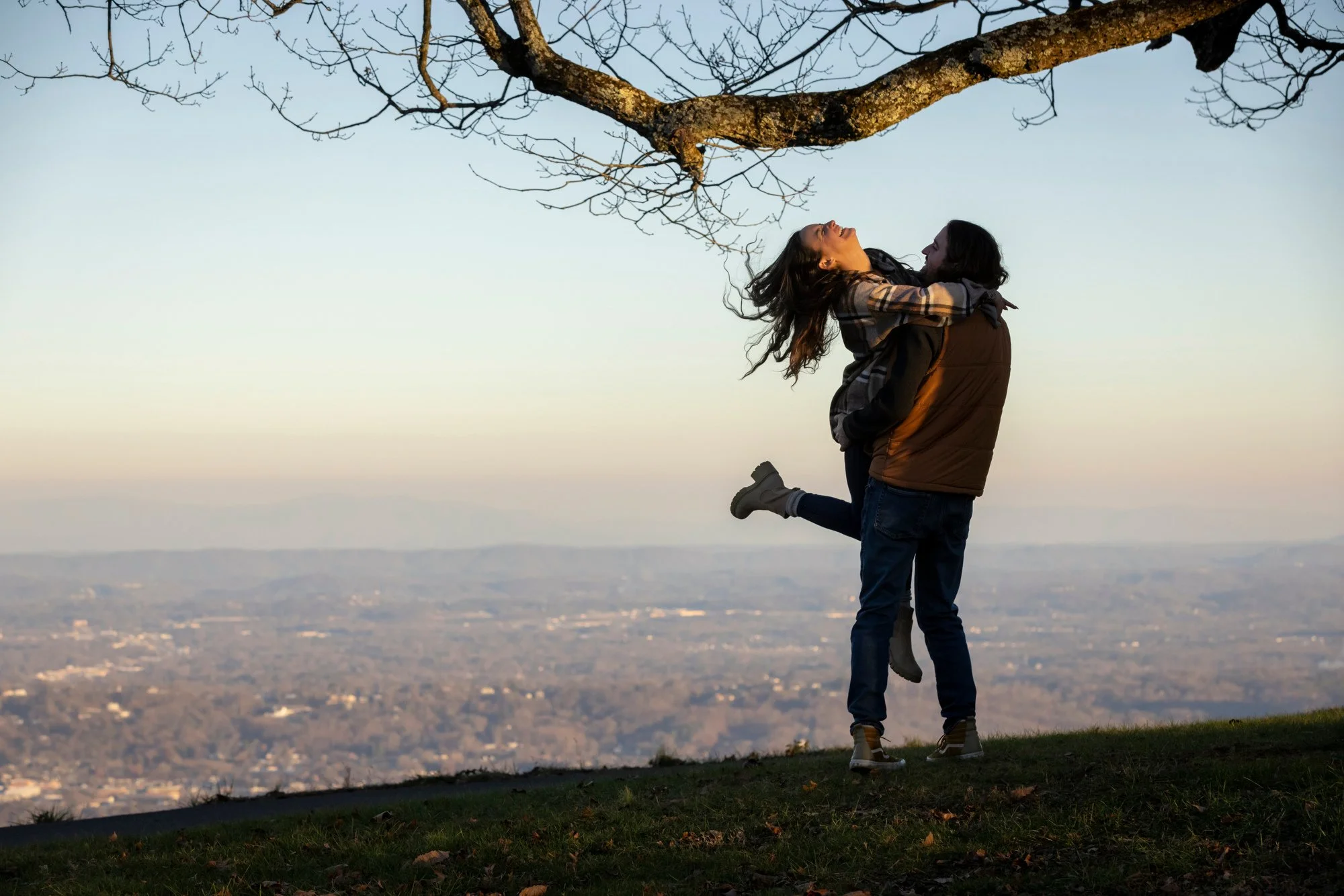 A couple sharing a joyful embrace and kiss, with the woman riding on the man's back, standing outdoors on a grassy hill with a scenic city view in the distance during sunset or sunrise.