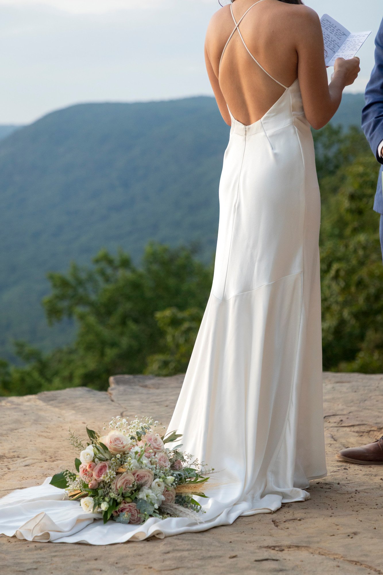 A woman in a white wedding dress standing outdoors near a mountain landscape, with a bouquet of pink and white roses on the ground nearby.