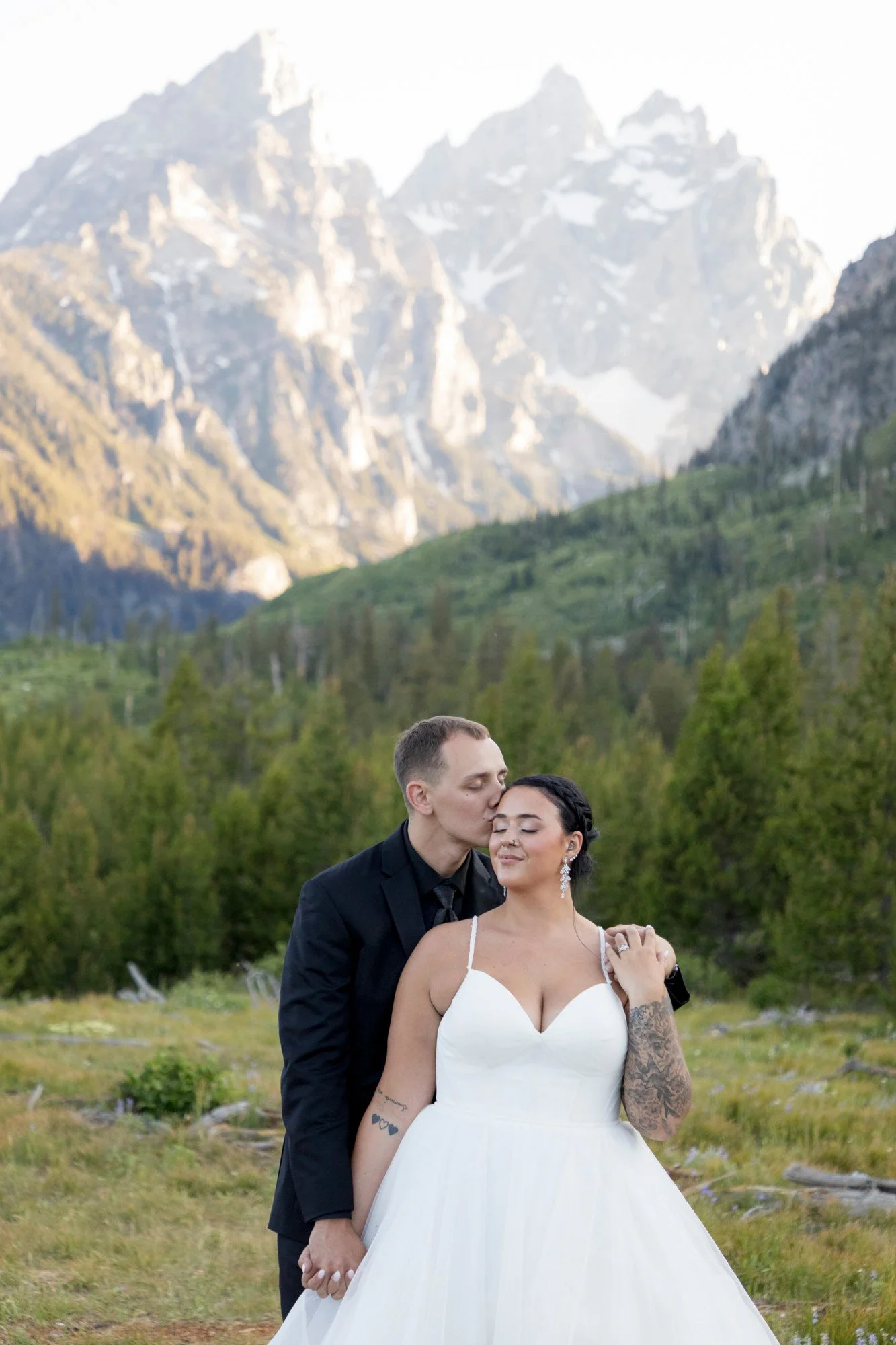 A newlywed couple, a man and a woman, standing close and holding hands outdoors with a mountainous landscape in the background. The man is kissing the woman on the forehead, and she is smiling with her eyes closed. The woman wears a white wedding dre