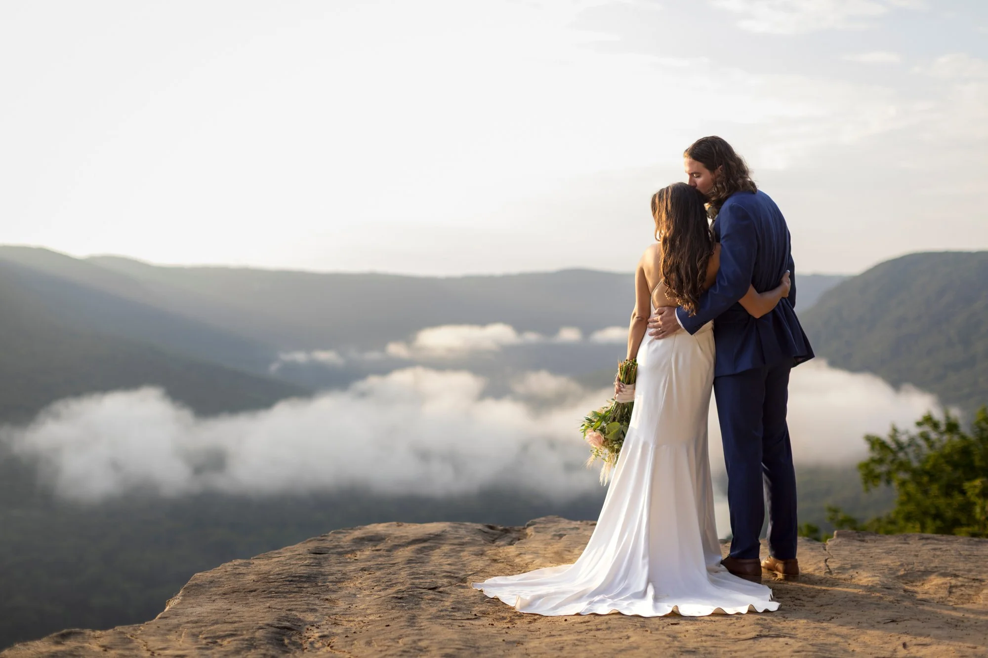 A bride and groom embrace on a cliff at sunset, with a scenic mountain view and cloudy sky in the background.