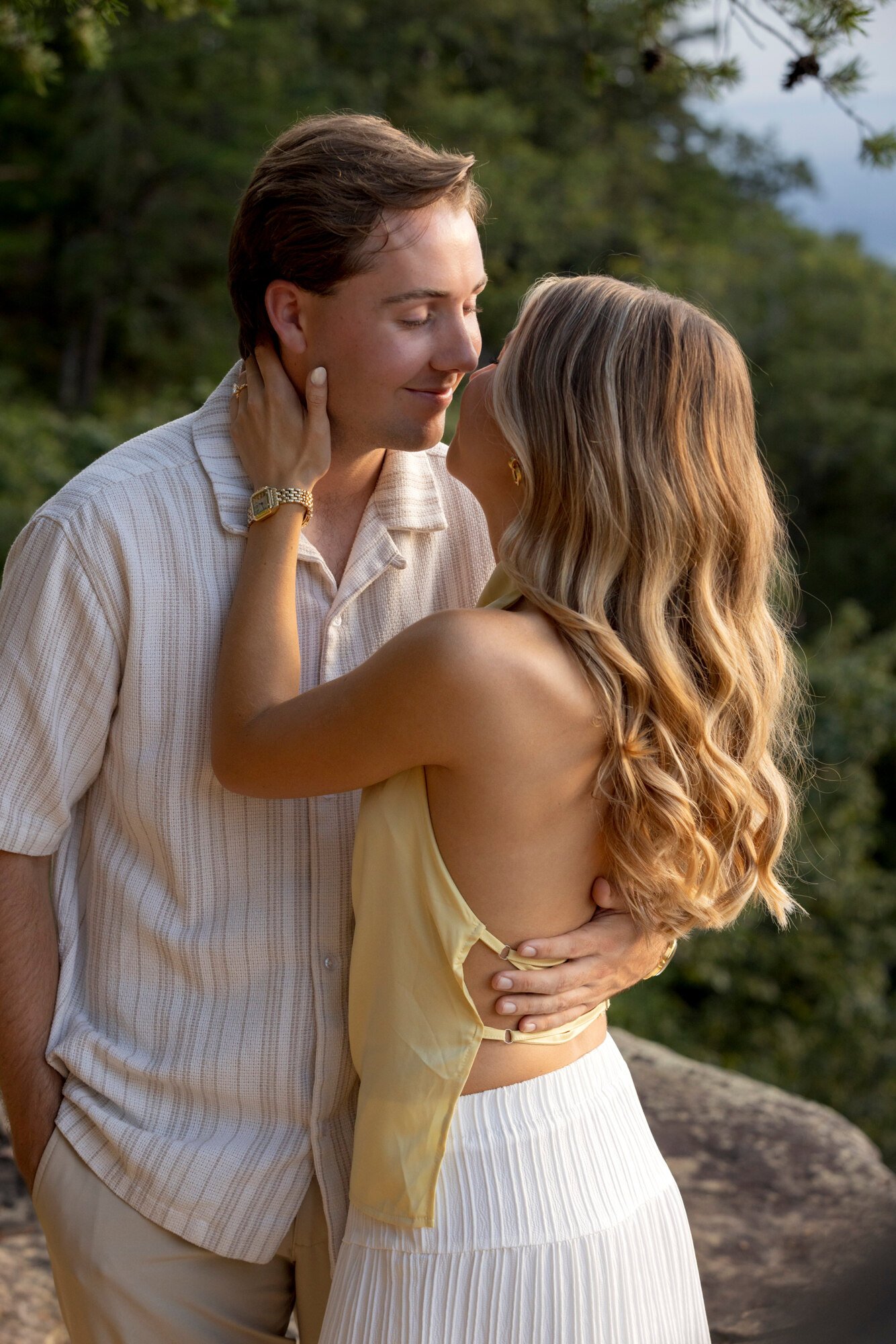 A couple is embracing outdoors, the woman is touching the man's face and they are about to kiss, with a background of trees and a cloudy sky.