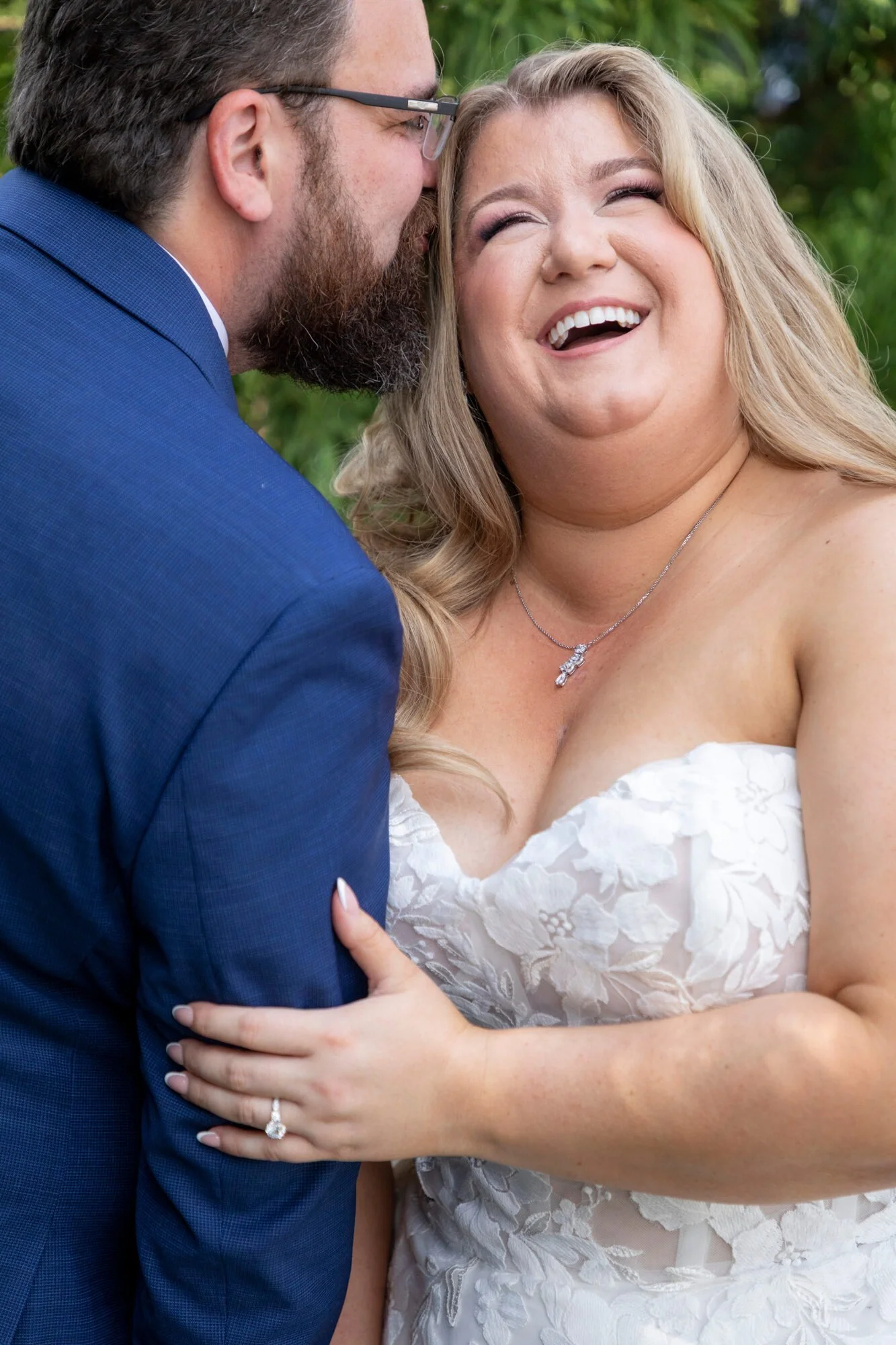Close-up of a smiling woman in a white wedding dress with a necklace, embracing a man dressed in a blue suit, outdoors with green foliage in the background.