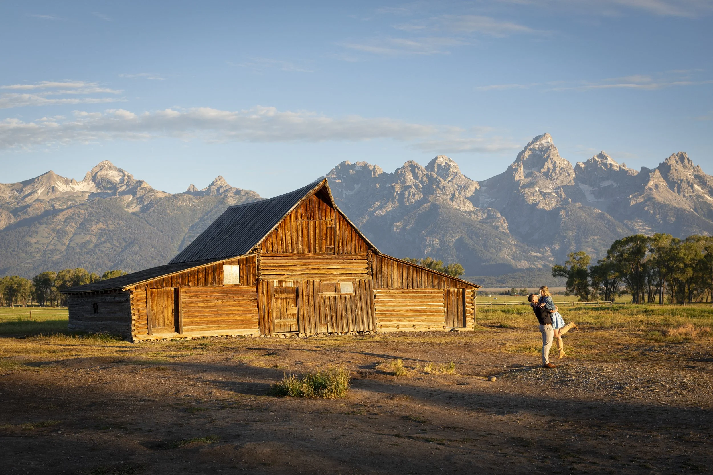 A wooden barn in front of a mountainous landscape with a person holding a child in a grassy field, during late afternoon or early evening sunlight.