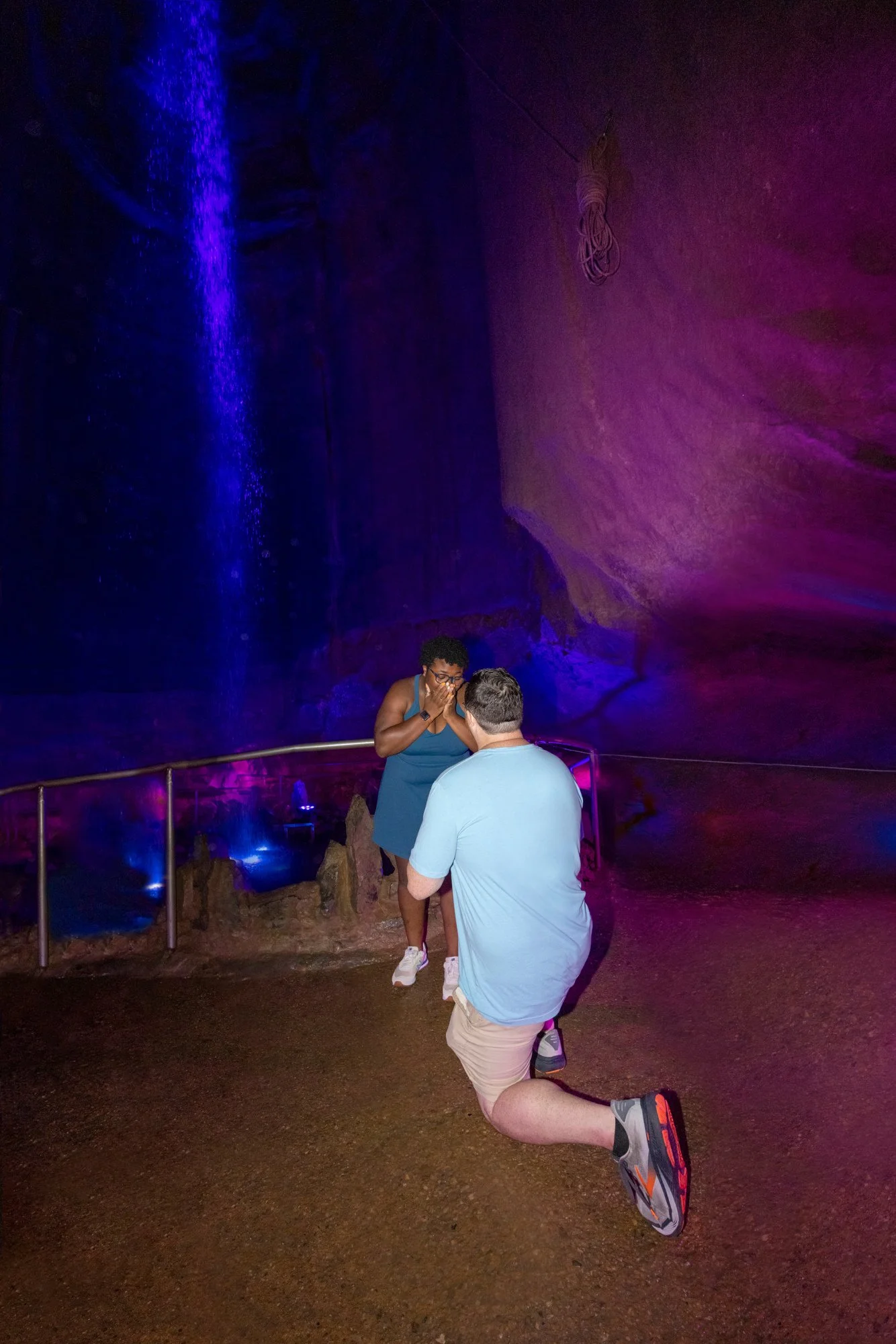 A person with a prosthetic leg kneeling on the ground, another person holding a ring, two people standing close to each other, and a cave-like background with purple and blue lighting.