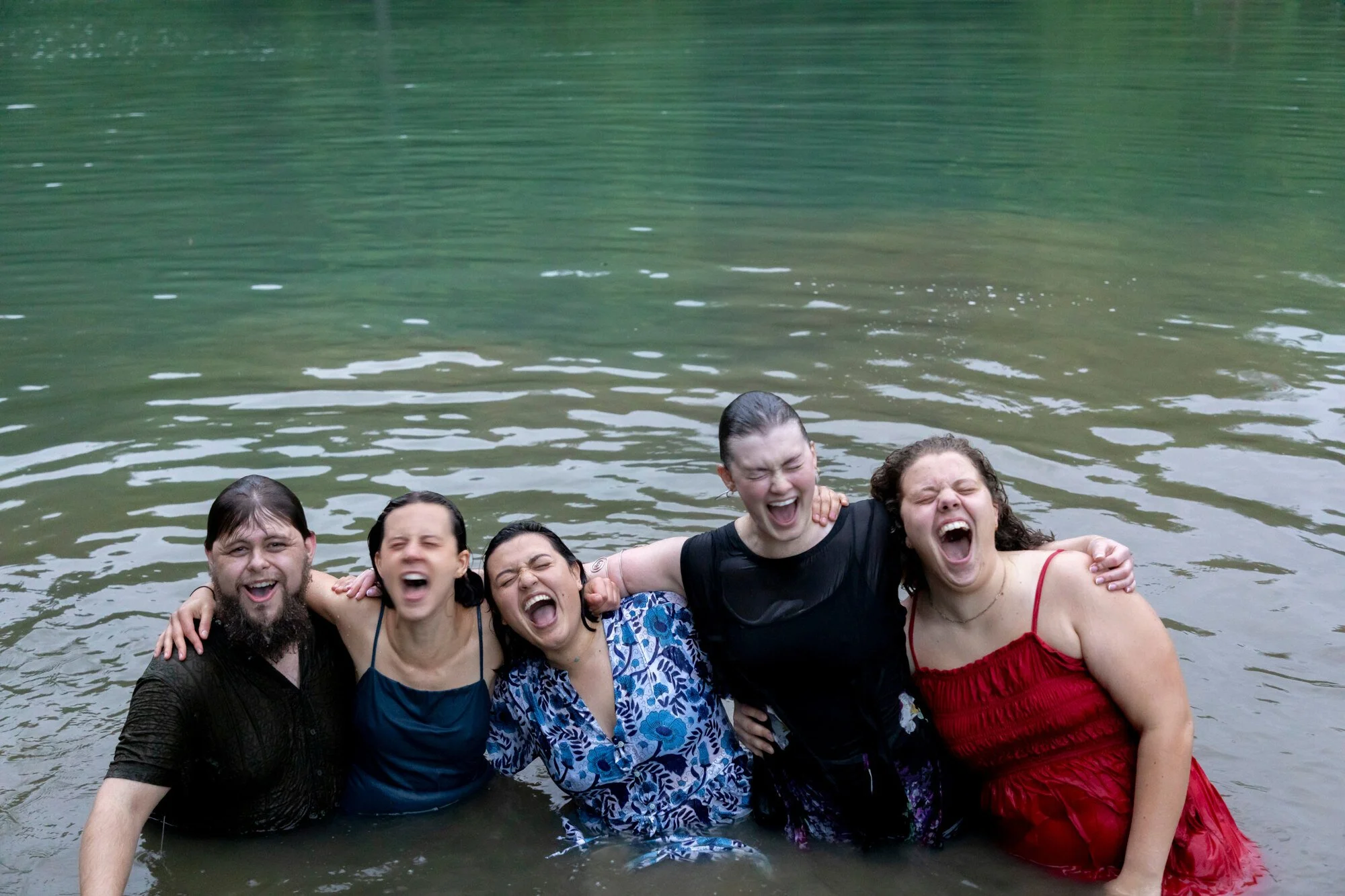 A group of six young adults are standing in water, smiling, and with outstretched arms, enjoying a swim. The background shows a greenish body of water.