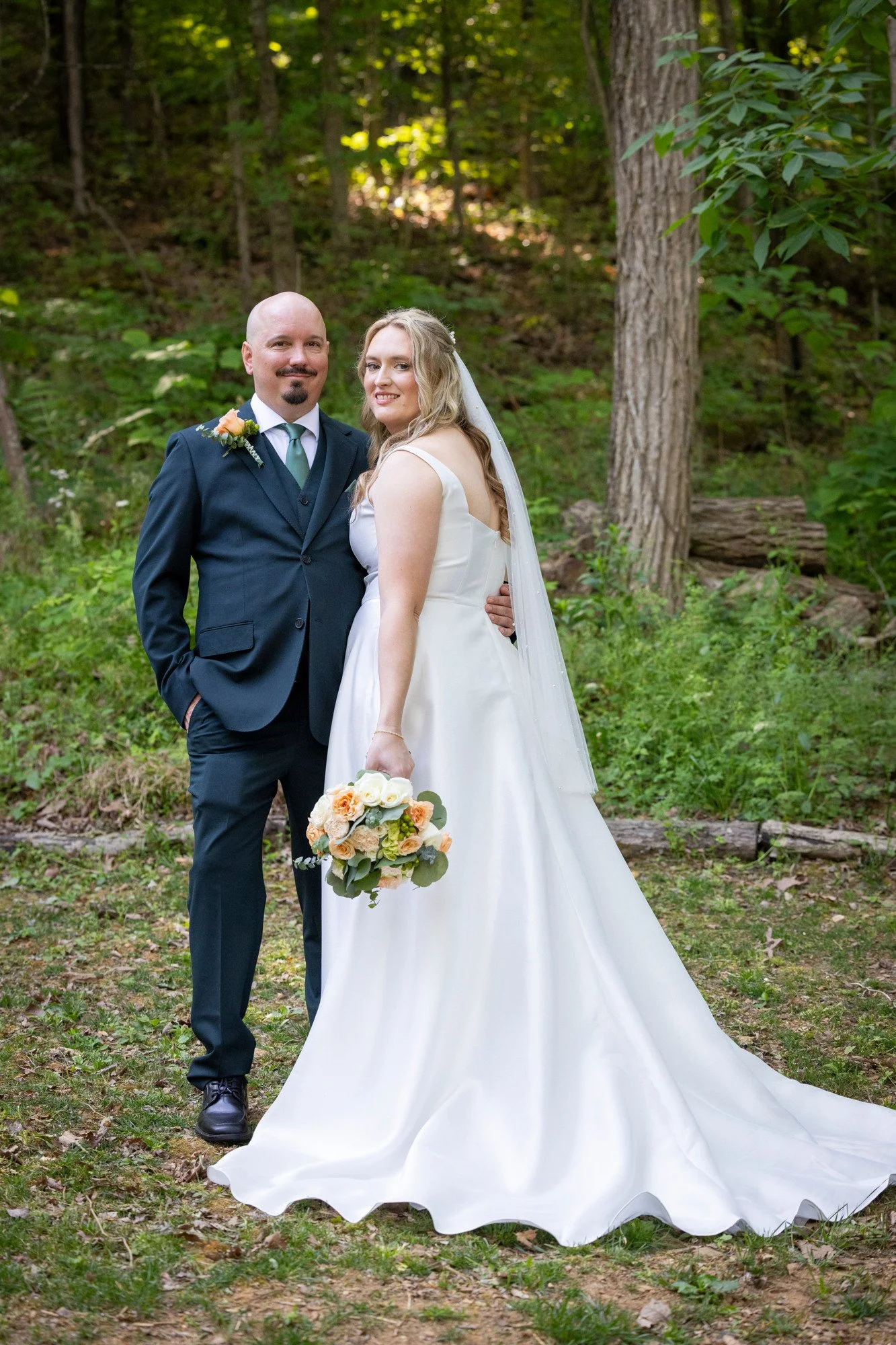 A newlywed couple stands in a wooded area for a wedding photo. The groom is wearing a navy suit with a light green tie and a peach boutonniere. The bride is in a white wedding gown, holding a bouquet of peach and cream flowers, and wearing a veil. Th
