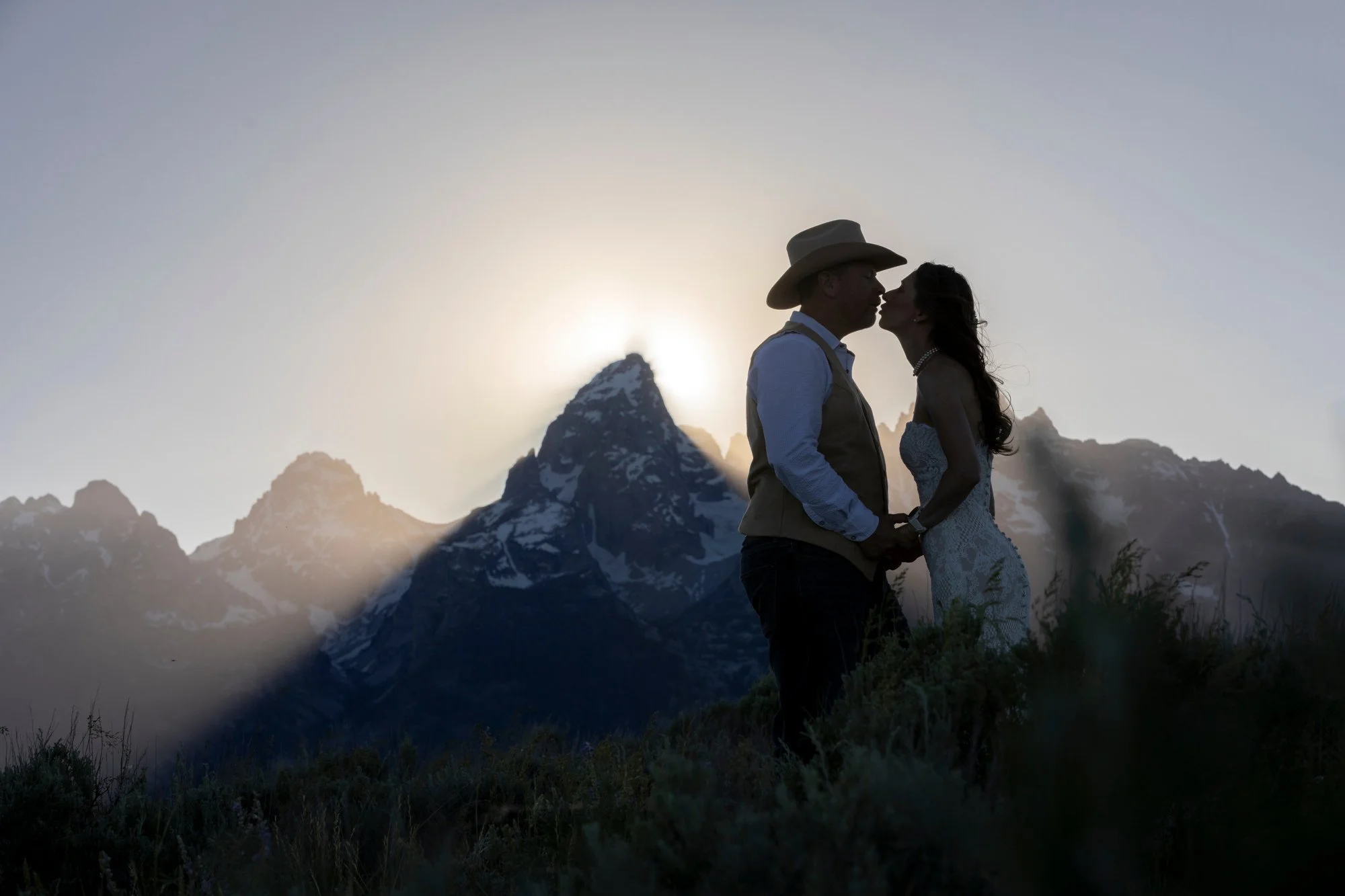 A silhouette of a couple kissing in front of a mountain range with a snow-capped peak, during sunset or sunrise.