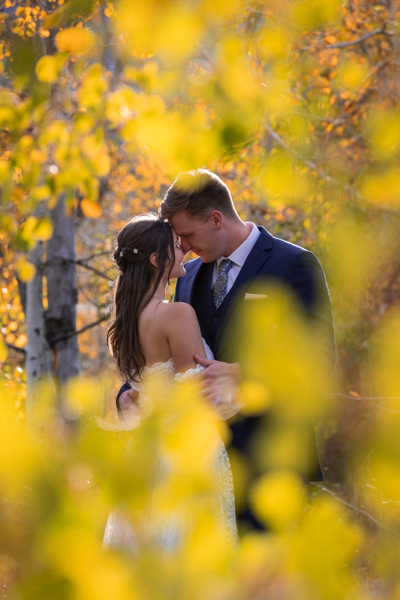 A couple, dressed in wedding attire, sharing a moment in a forest with autumn yellow leaves.